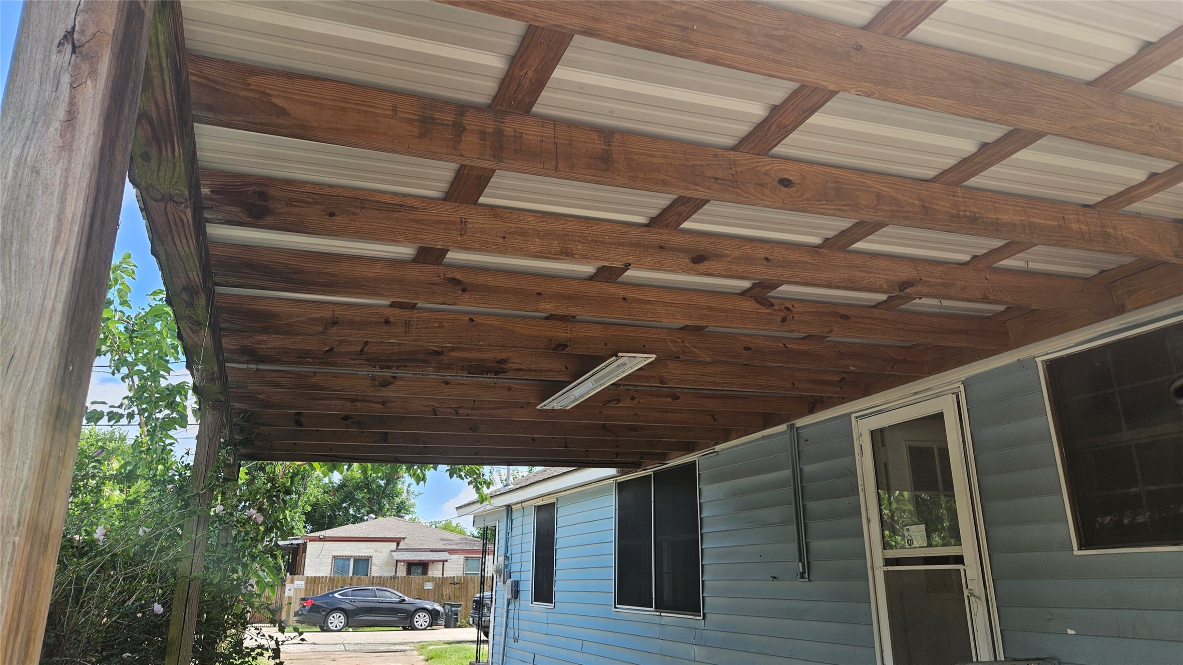 2415 Perry Street Baytown, TX 77521 - Photo 18 of 23 This photo shows a covered patio area with a wooden frame and a metal roof, attached to a blue-sided house. It's a simple outdoor space, potentially useful for relaxation or gatherings.