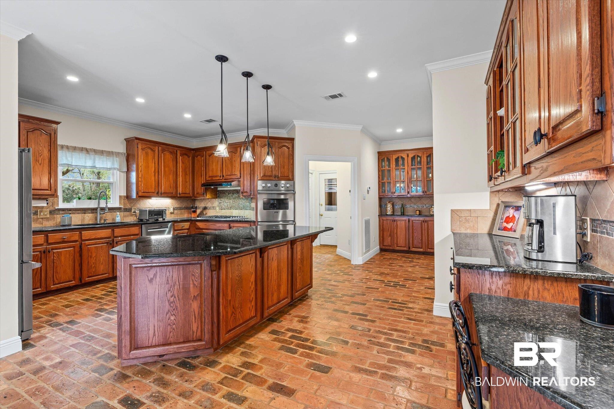 4851 Audubon Drive Mobile, AL 36619 - Photo 8 of 46 Kitchen featuring brown cabinetry, dark stone countertops, glass insert cabinets, ornamental molding, and brick flooring