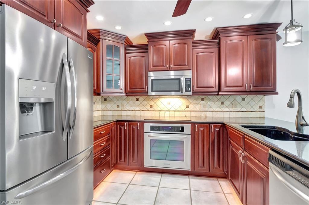 3035 42nd Terrace Southwest Naples, FL 34116 - Photo 17 of 48 Kitchen with stainless steel appliances, bold wood finish cabinets, dark stone countertops, glass fronted cabinets, and decorative light fixtures.