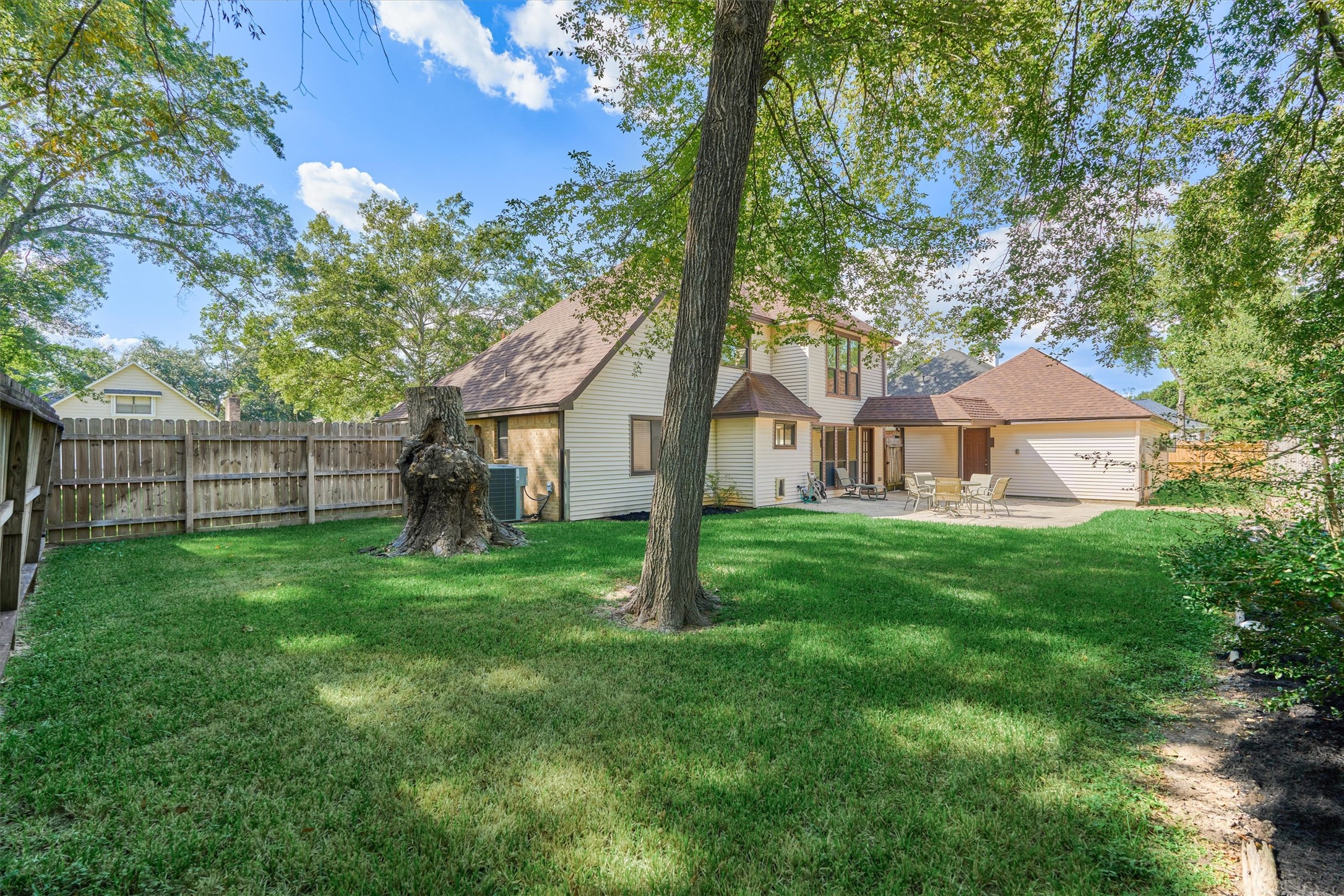 25706 Overlake Drive Spring, TX 77380 - Photo 30 of 32 Another view of the spacious backyard with lush green grass and mature trees, providing a serene and private outdoor space. The house features a light exterior with a fenced perimeter, an outdoor seating area, and a two-story structure with a sloped roof, enhancing its charm and appeal.