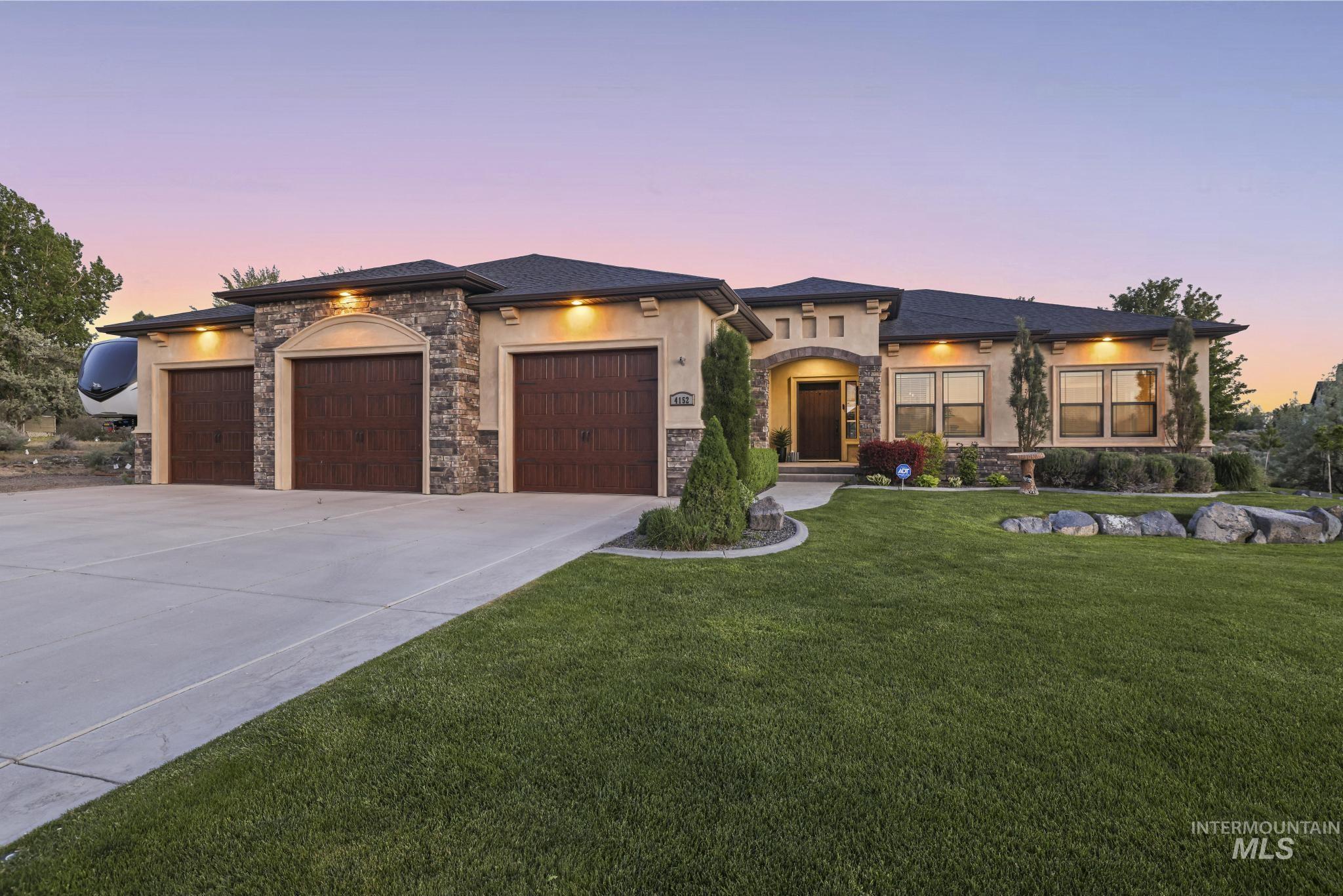 4152 North Meadow Ridge Circle Twin Falls, ID 83301 - Photo 1 of 50 Stucco and stone exterior featuring a three-car garage with wood-finish doors, a concrete driveway, a manicured lawn, and mature landscaping