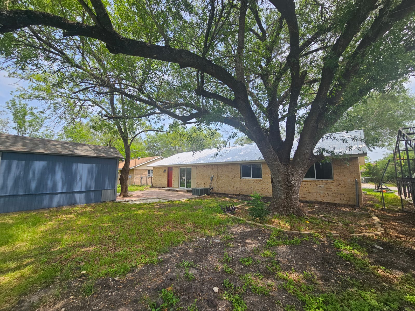 1401 Trinity Street Lockhart, TX 78644 - Photo 29 of 31 Expansive backyard featuring mature shade trees, a brick exterior, sliding glass door access, a blue-sided utility building, and a metal roof