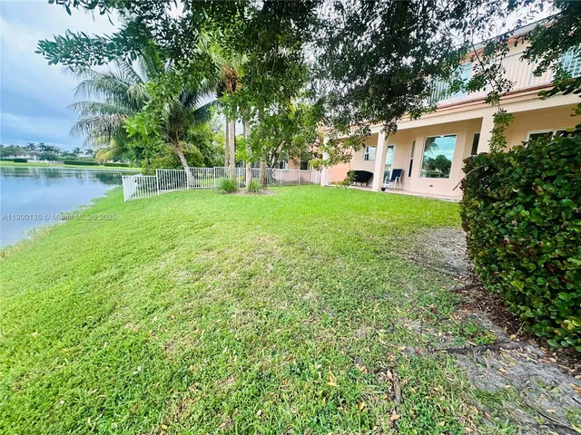 a view of a white house in front of a big yard with plants and large trees