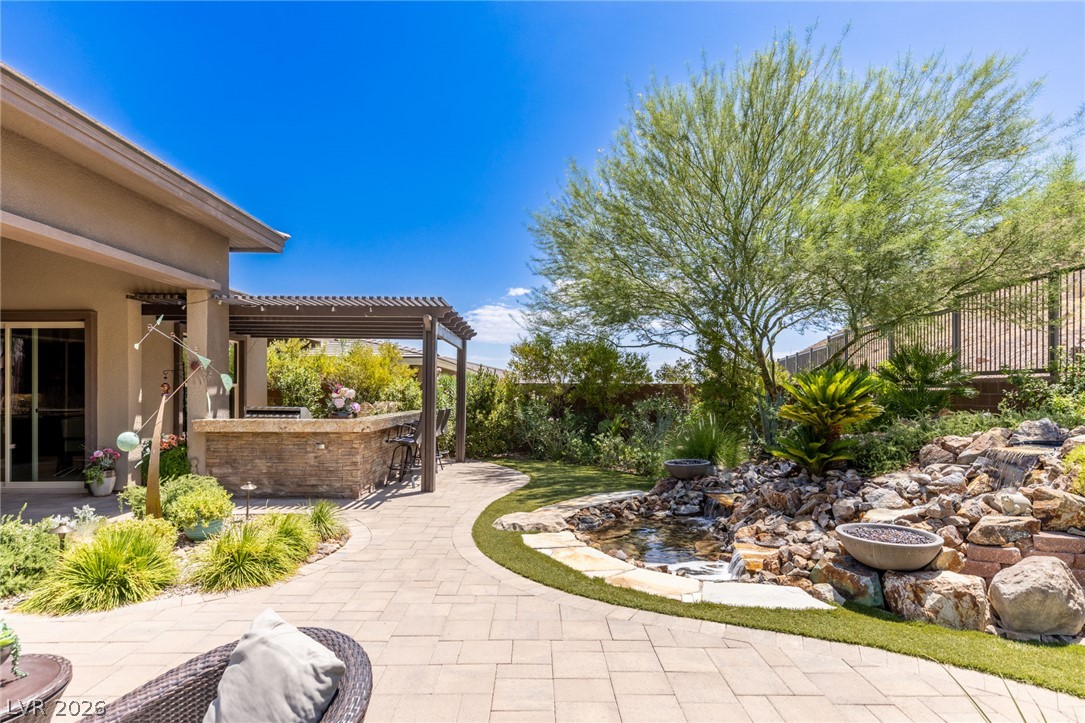 10167 Copper Edge Road Las Vegas, NV 89148 - Photo 34 of 45 A private backyard oasis features an outdoor kitchen beneath a pergola, complemented by a rock-lined pond and fire bowls.
