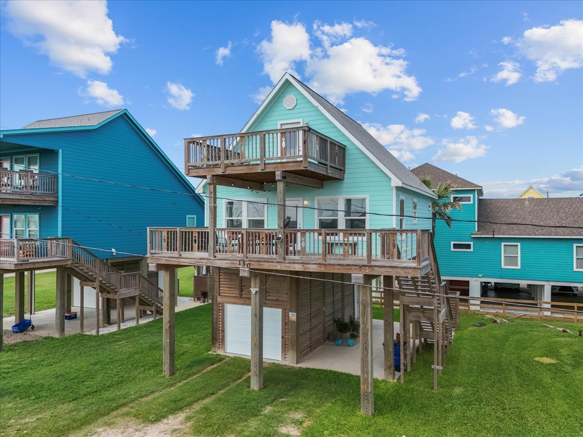 130 Beachcomber Avenue Surfside Beach, TX 77541 - Photo 1 of 33 This charming elevated home features a bright sea glass teal exterior with white trim, multiple wooden decks for outdoor enjoyment, and is supported by stilts, typical for coastal properties. The surrounding area is grassy and open, providing a tranquil setting.