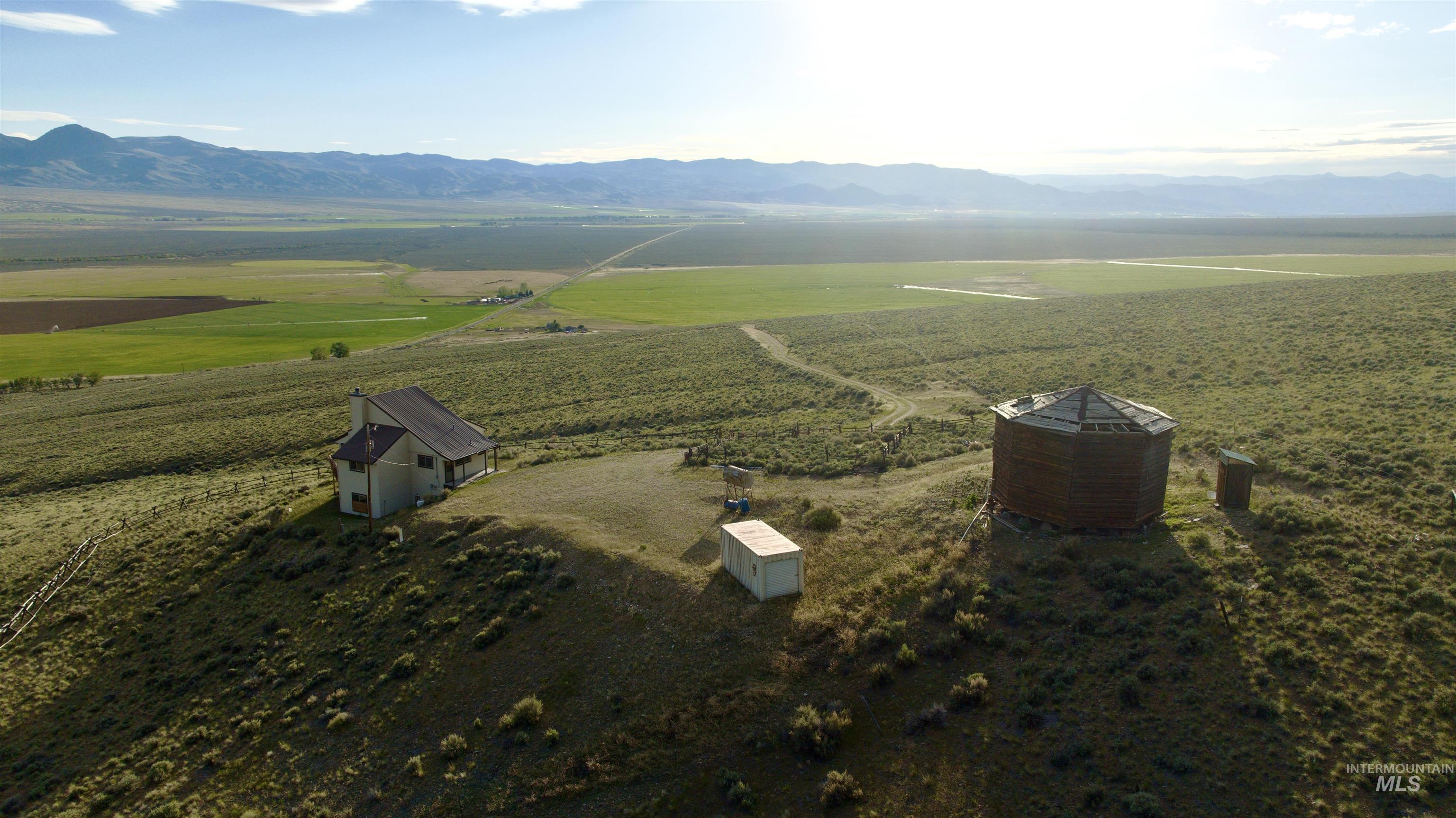 2040 Pahsimeroi River Road May, ID 83253 - Photo 8 of 29 Rural property featuring a two-story residence with a dark-pitched roof, a large octagonal outbuilding, and a small utility structure, all surrounded by expansive natural landscaping with distant agricultural fields and mountain ranges