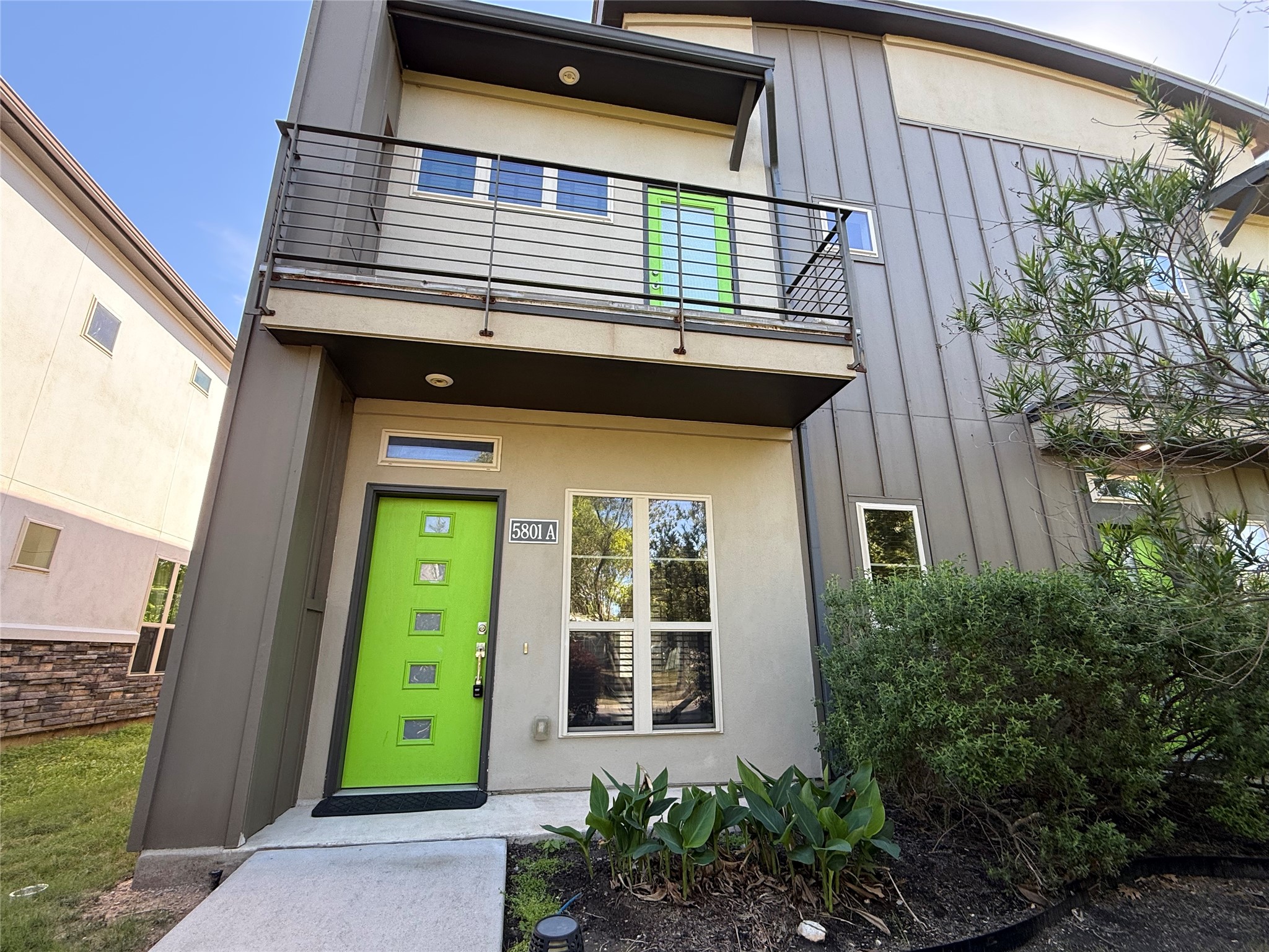 Modern exterior with a vibrant green front door, a second-story balcony, and a shaded landscaped front yard.