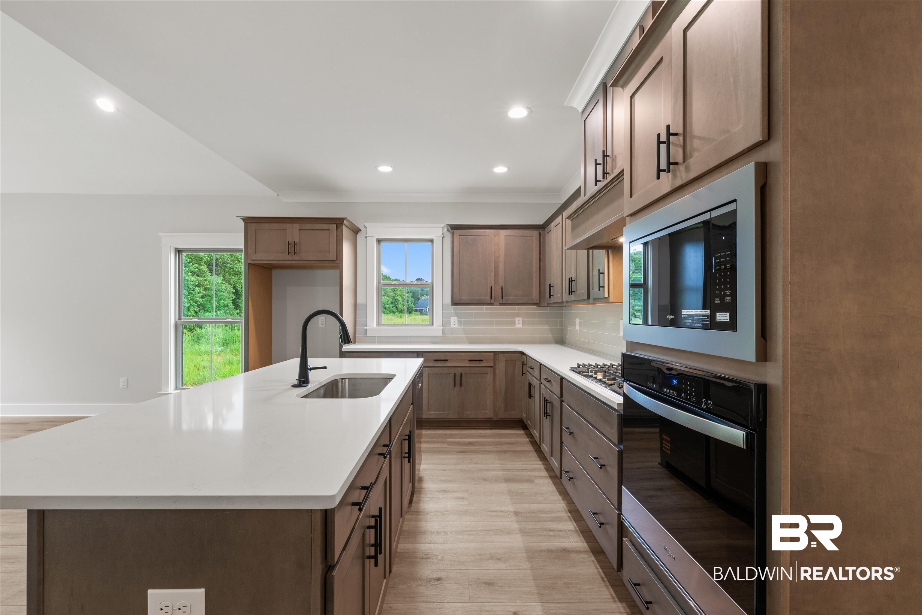 27494 Patch Pl Loop Daphne, AL 36526 - Photo 10 of 26 Modern kitchen featuring an expansive island with a stainless steel sink and matte black faucet, custom cabinetry with dark hardware, light wood-finish flooring, and integrated stainless steel appliances including a wall oven and microwave