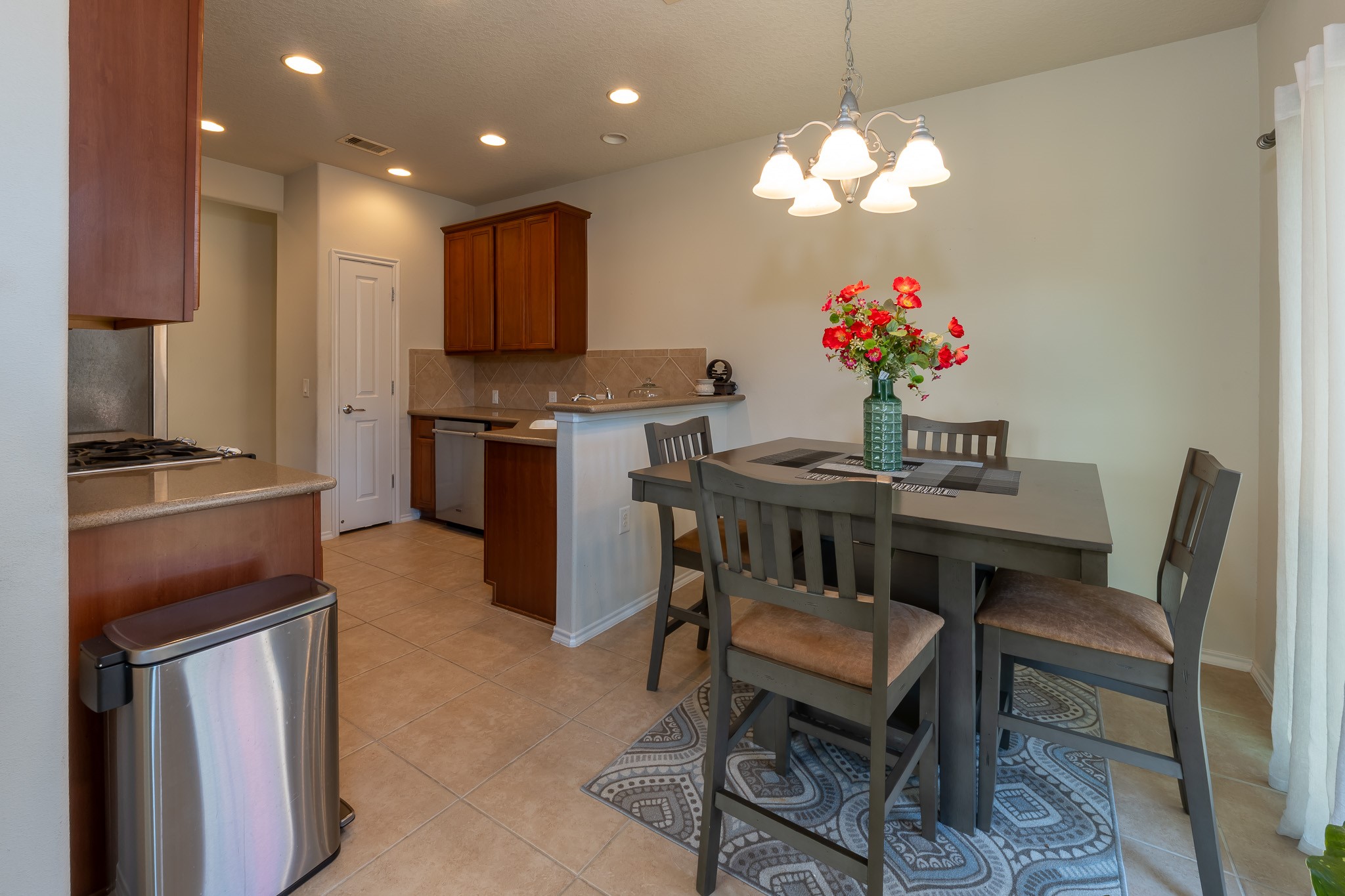 13600 Breton Ridge Street, Unit 29F Houston, TX 77070 - Photo 8 of 21 This photo shows a cozy dining area with a four-seater table and a decorative centerpiece, adjacent to a kitchen featuring wooden cabinets, a dishwasher, and ample counter space. The space is well-lit with modern fixtures, and the neutral tile flooring complements the warm tones of the wood.