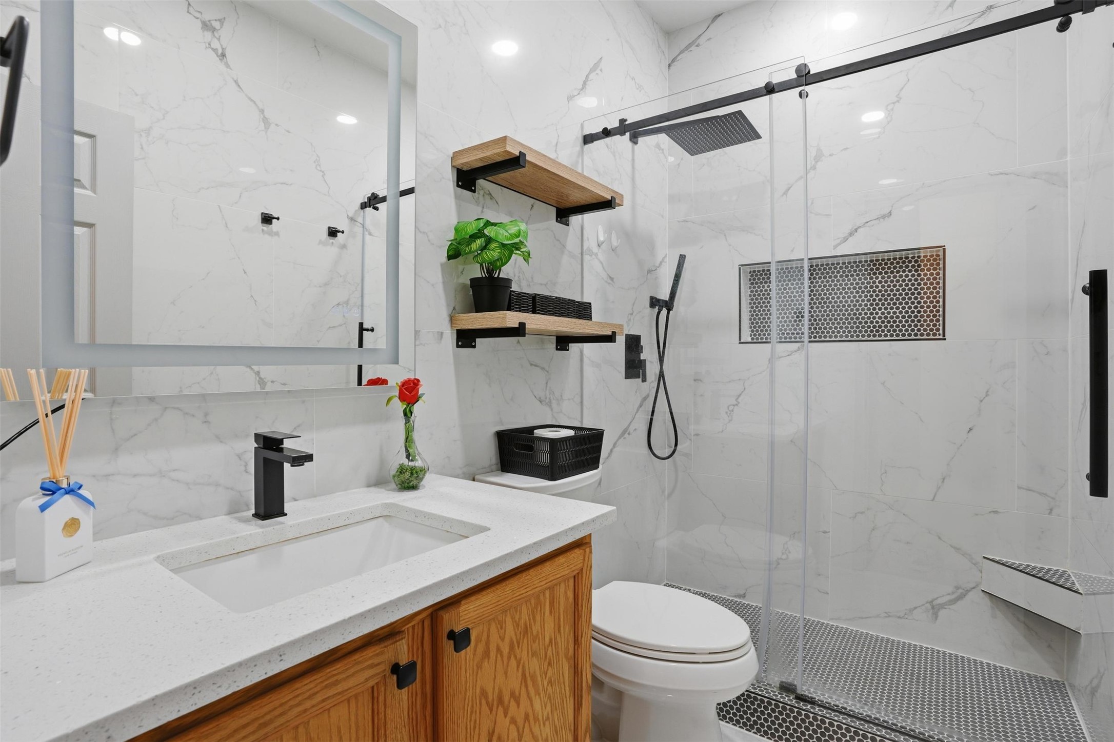 11606 Murchison Street Manor, TX 78653 - Photo 26 of 35 Secondary Bathroom features a vanity with a white countertop and a matte black faucet, a large mirror with an illuminated frame, and a shower with a sliding glass door and a built-in shelf. Wall to ceiling marble-like tile.
