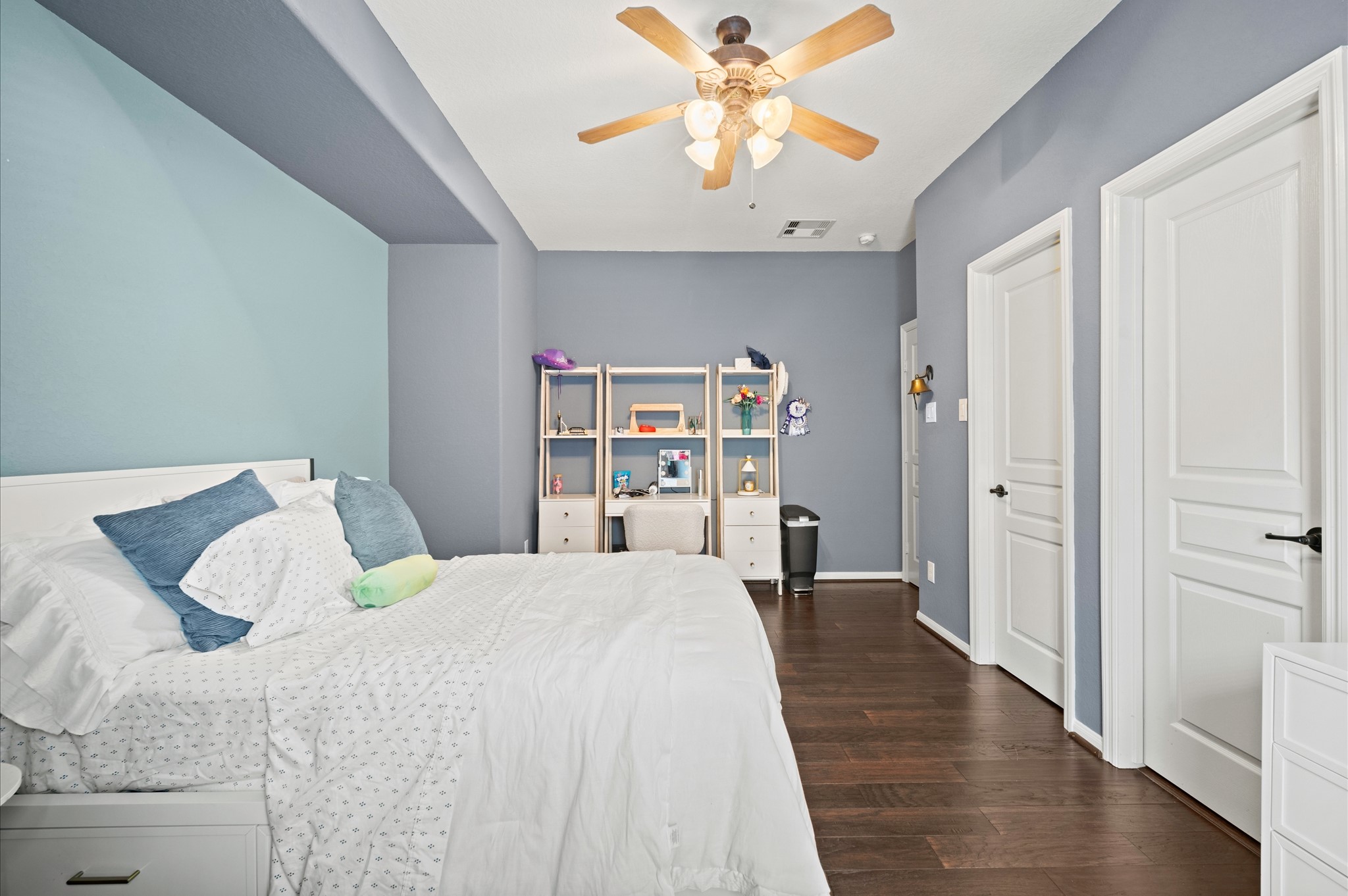 9507 Empress Crossing Drive Spring, TX 77379 - Photo 32 of 50 This bedroom features a calming blue and gray color scheme with hardwood floors. It includes a ceiling fan, ample natural light, and a cozy bed. A built-in shelf provides storage and display space, and there are two doors for easy access.