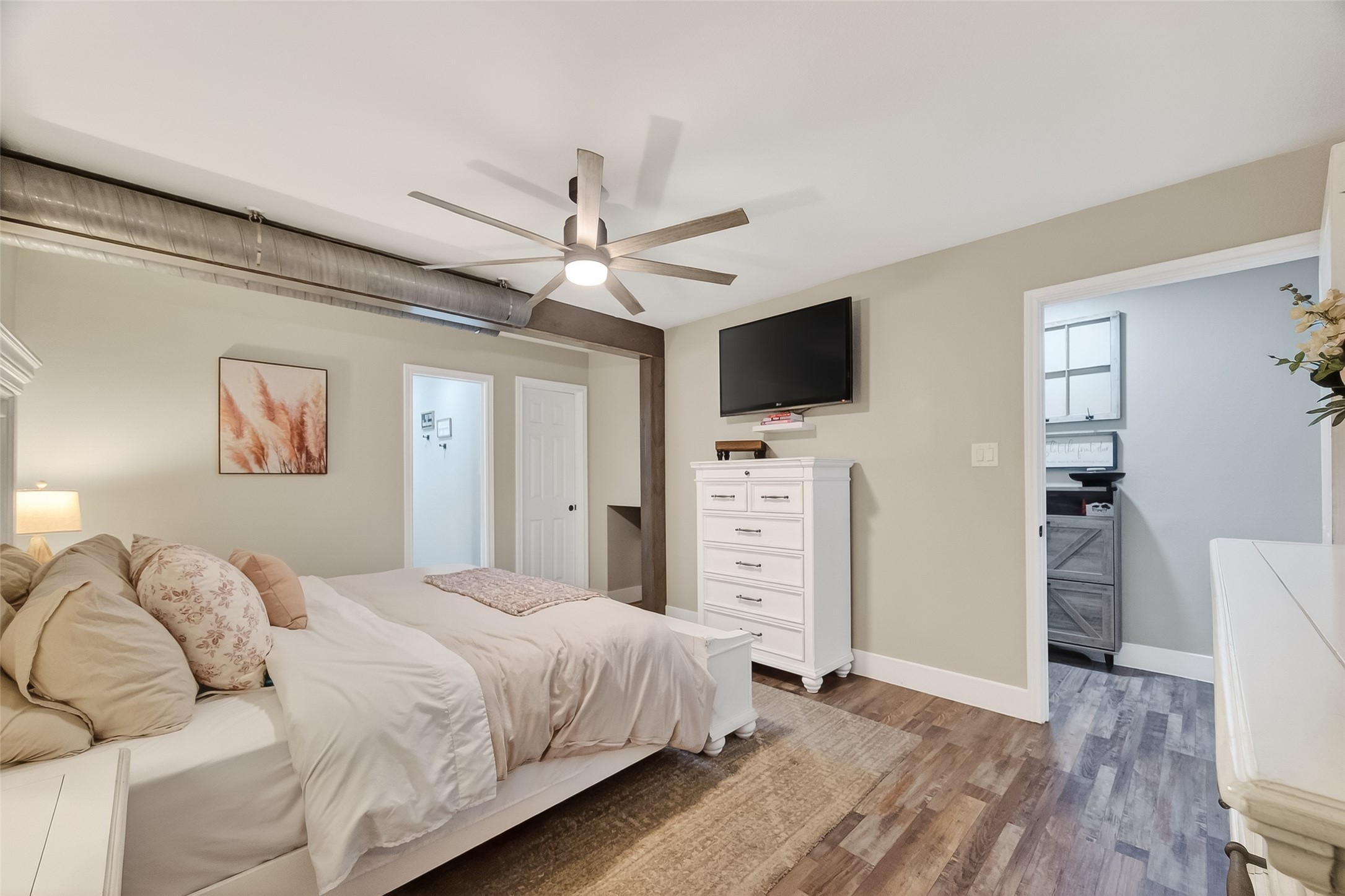 819 Sentinel Oaks Pinehurst, TX 77362 - Photo 21 of 38 This bedroom features a cozy design with neutral tones and wood flooring. It includes a modern ceiling fan, wall-mounted TV, and ample storage with a tall dresser. The room connects to an adjacent space through open doorways, adding convenience.