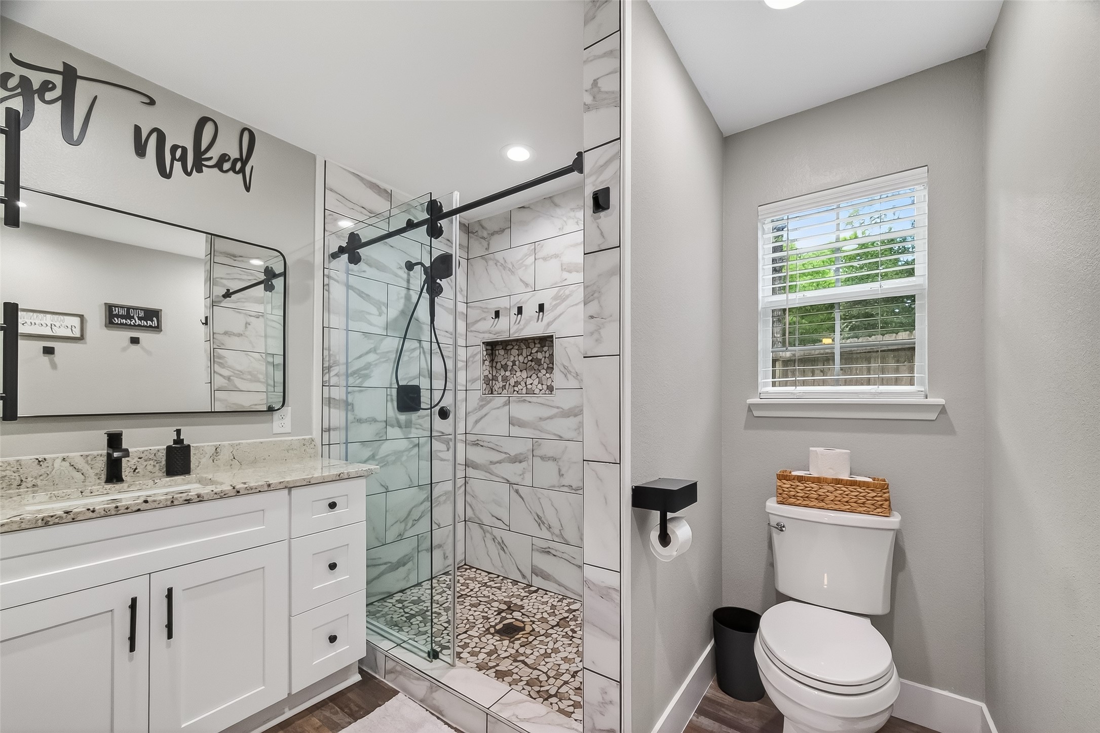 819 Sentinel Oaks Pinehurst, TX 77362 - Photo 23 of 38 Modern bathroom with a spacious glass-enclosed shower featuring marble tile and pebble flooring. It has a sleek white vanity with granite countertops, stylish black fixtures, and a large mirror. Natural light enters through a window above the toilet.