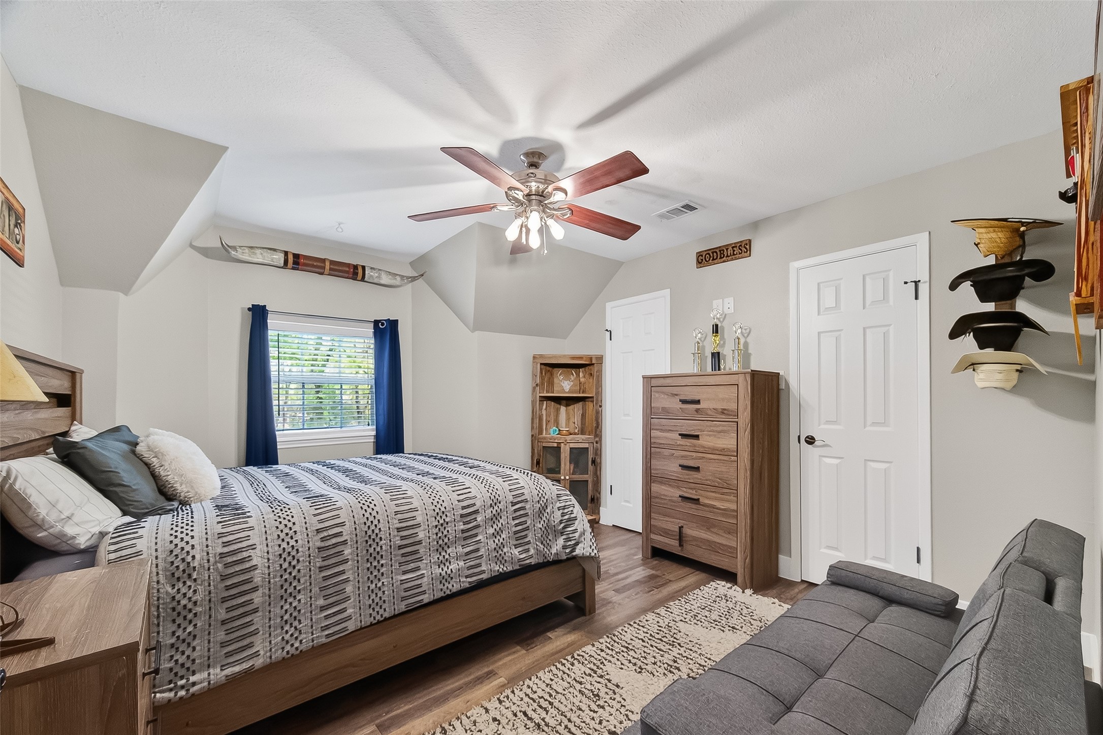 819 Sentinel Oaks Pinehurst, TX 77362 - Photo 25 of 38 This cozy bedroom features a sloped ceiling with a fan, a comfortable bed, and modern wooden furniture. A window with blue curtains provides natural light, and the room is accented with decorative elements, offering a welcoming and functional space.