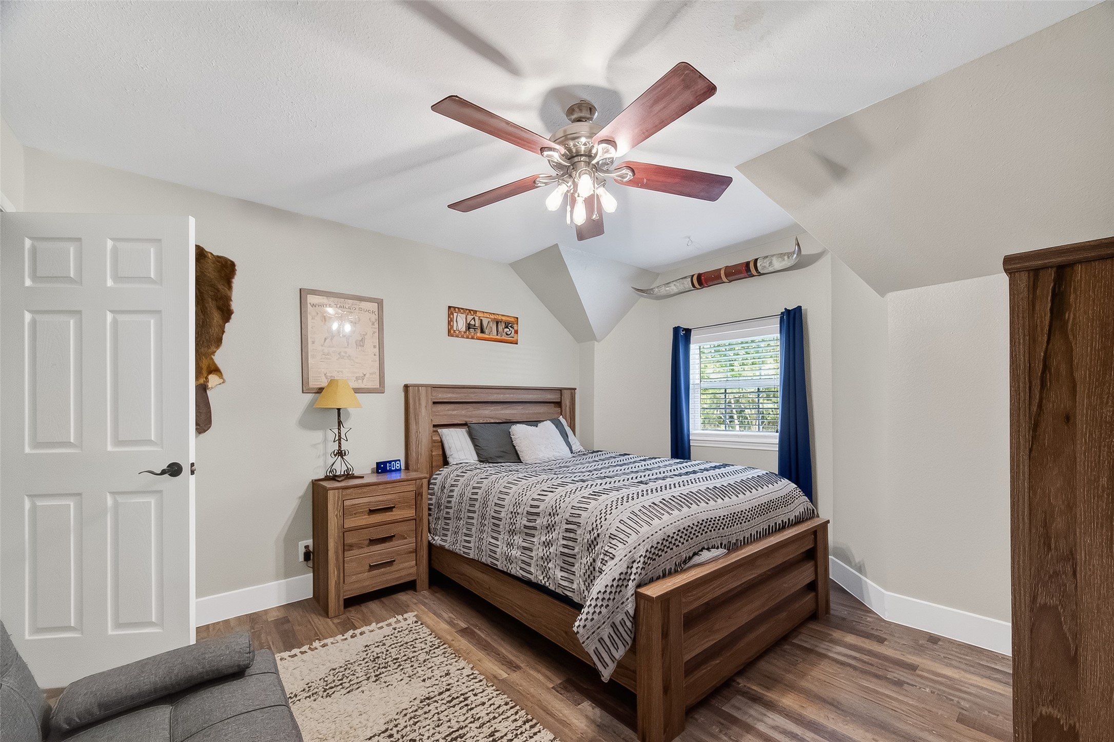 819 Sentinel Oaks Pinehurst, TX 77362 - Photo 26 of 38 This cozy bedroom features a wooden floor and bed, a ceiling fan, and a window with blue curtains providing natural light. The room is decorated with a wall-mounted art piece and a small nightstand with a lamp.