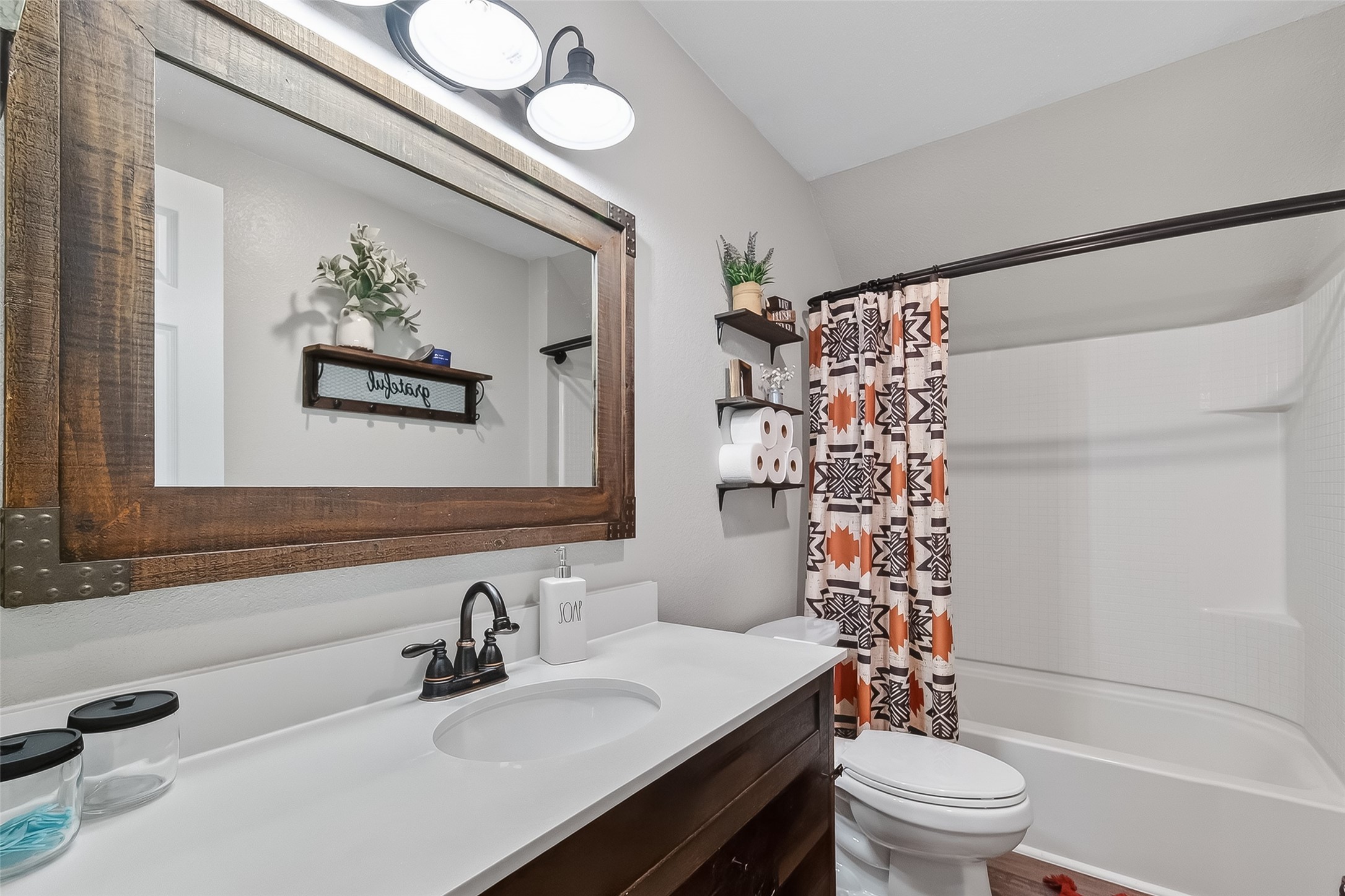 819 Sentinel Oaks Pinehurst, TX 77362 - Photo 27 of 38 This bathroom features a rustic wooden mirror, sleek vanity with black fixtures, and a shower-tub combo. The space is accented with a patterned shower curtain and open shelving for storage.