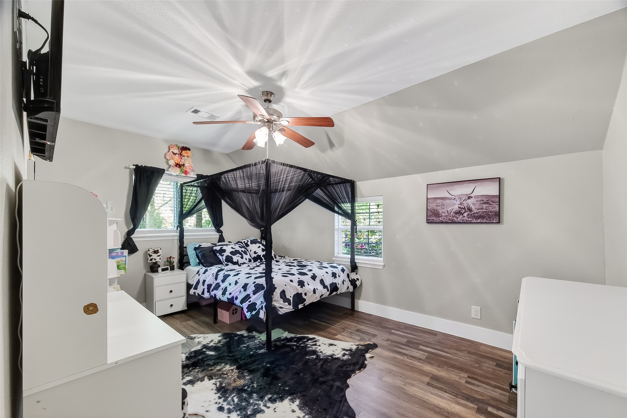 819 Sentinel Oaks Pinehurst, TX 77362 - Photo 28 of 38 This bedroom features a cozy atmosphere with hardwood flooring and neutral walls. It includes a bed with a canopy, a ceiling fan, and two windows for natural light. The room is accented with a cowhide rug and wall art, offering a blend of comfort and style.