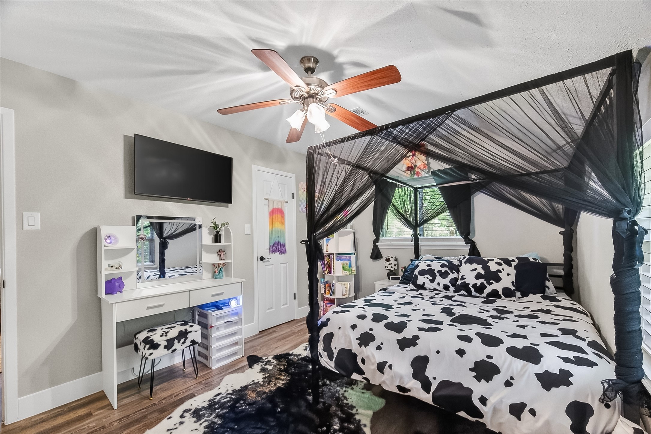 819 Sentinel Oaks Pinehurst, TX 77362 - Photo 29 of 38 This bedroom features a stylish canopy bed with cow-print bedding, a matching rug, and a ceiling fan. It includes a vanity with a mirror, a mounted TV, and ample natural light from large windows, creating a cozy and inviting space.