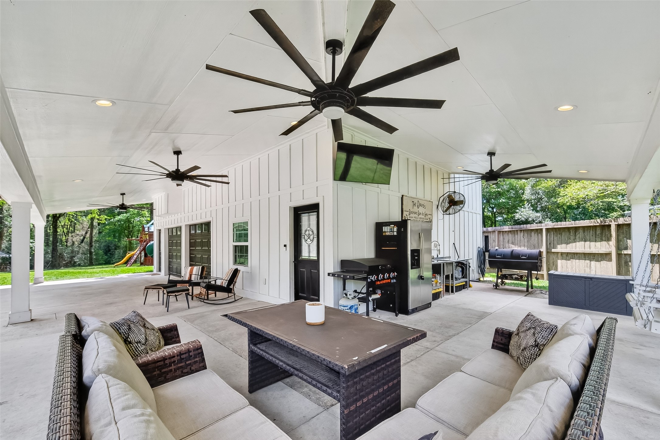 819 Sentinel Oaks Pinehurst, TX 77362 - Photo 7 of 38 This photo showcases a spacious covered patio with ceiling fans, comfortable seating, and an outdoor kitchen area, perfect for entertaining. The area overlooks a green backyard with a fence and a playset, offering a blend of relaxation and family-friendly features.