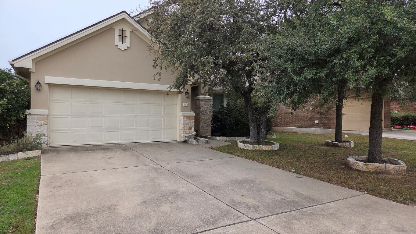 View of property hidden behind natural elements with stucco siding, concrete driveway, stone siding, and a front yard