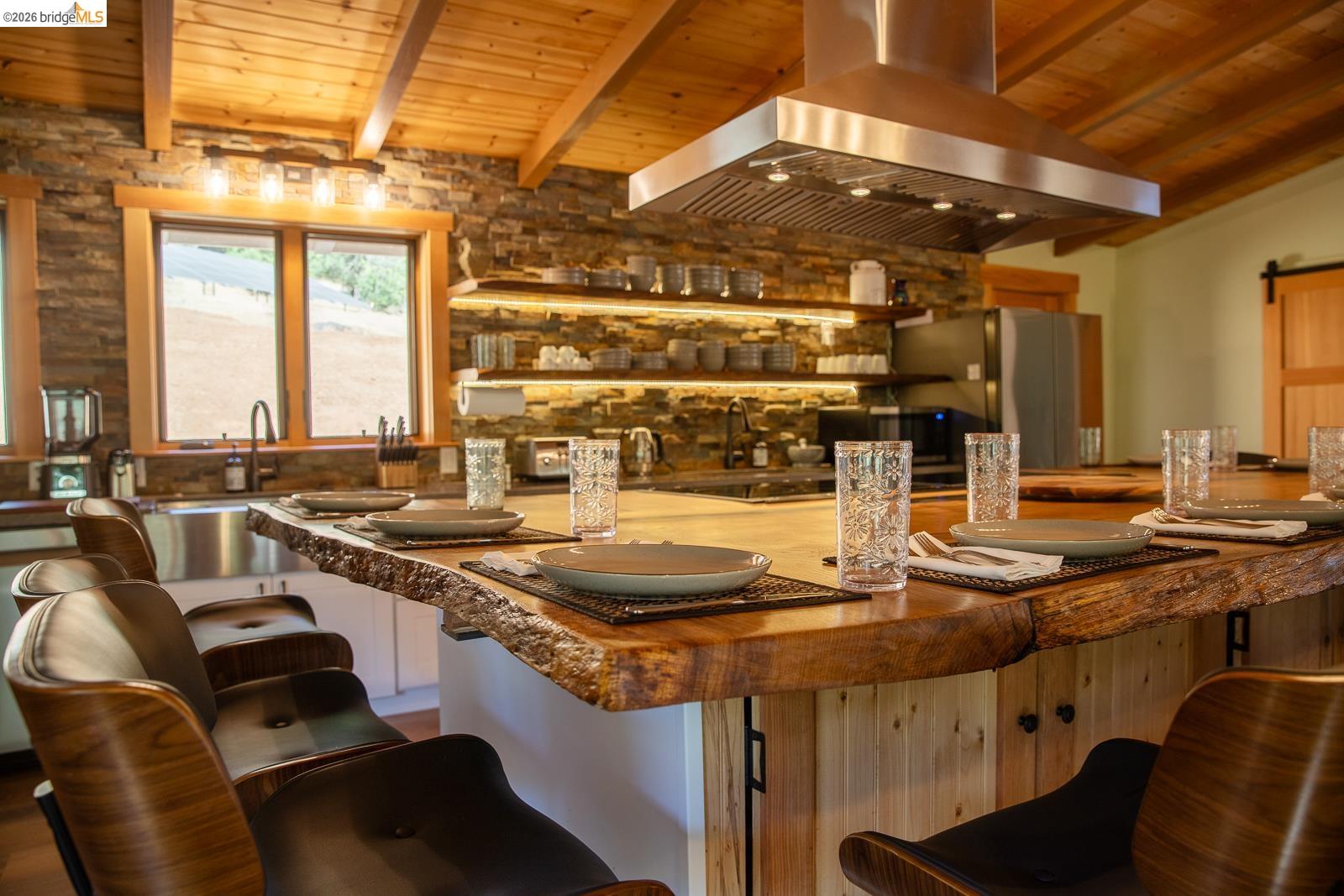 18399 Lime Kiln Road Sonora, CA 95370 - Photo 7 of 36 Kitchen featuring a live-edge wood island, exposed wood beam ceiling, stacked stone accent wall, recessed under-shelf lighting, and a stainless steel range hood