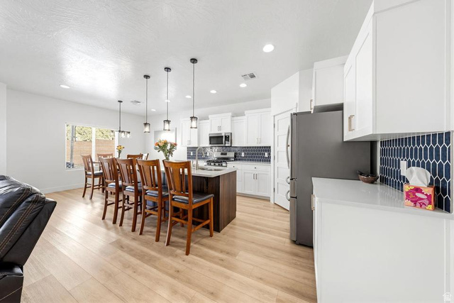 5963 Flattop Drive St. George, UT 84790 - Photo 10 of 45 Bright kitchen featuring white cabinetry, light wood-finish flooring, a dark-toned island with pendant lighting, a patterned blue backsplash, and stainless steel appliances