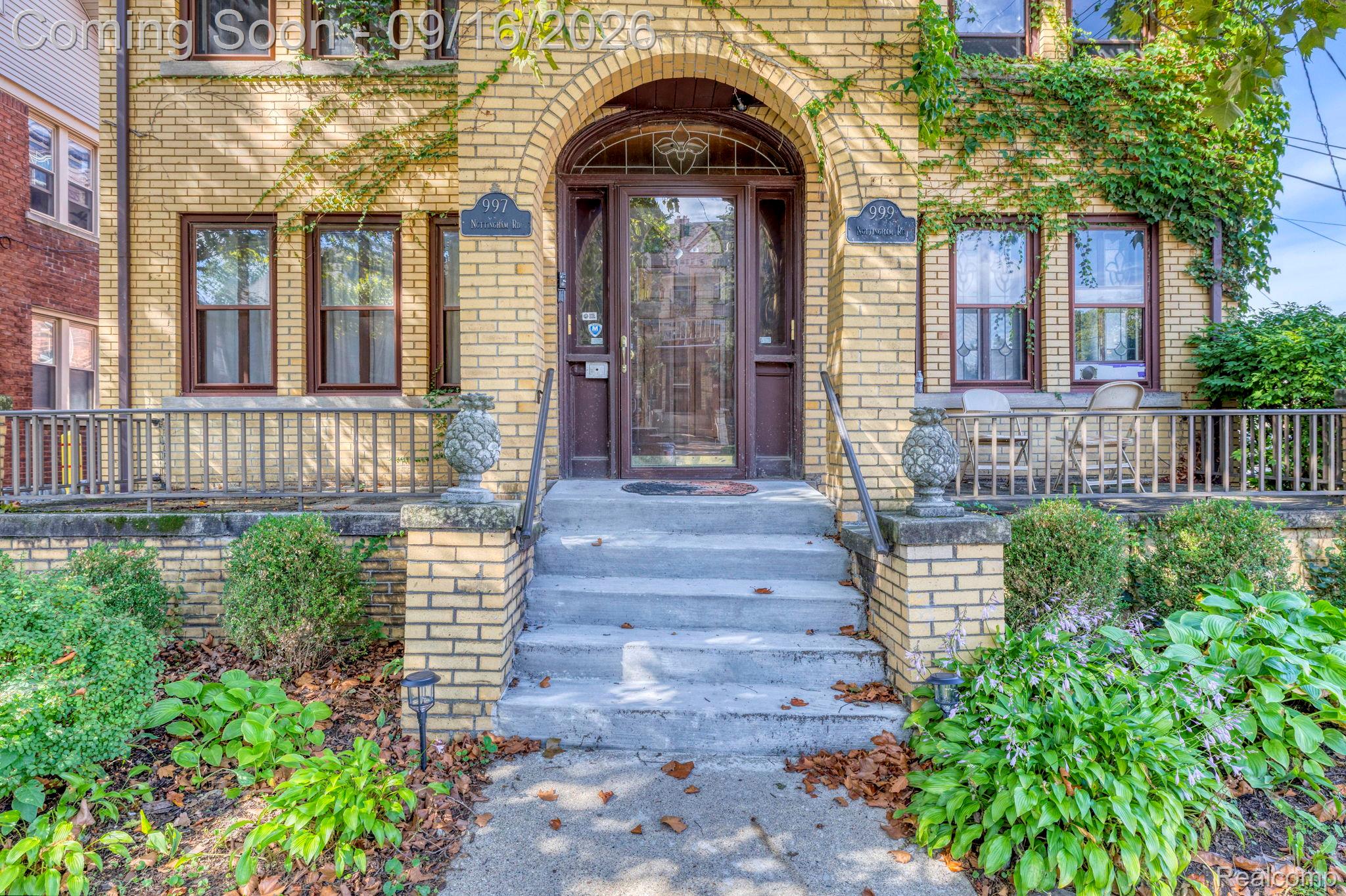 Brick facade with an arched entry, covered porch, concrete steps, and mature landscaping