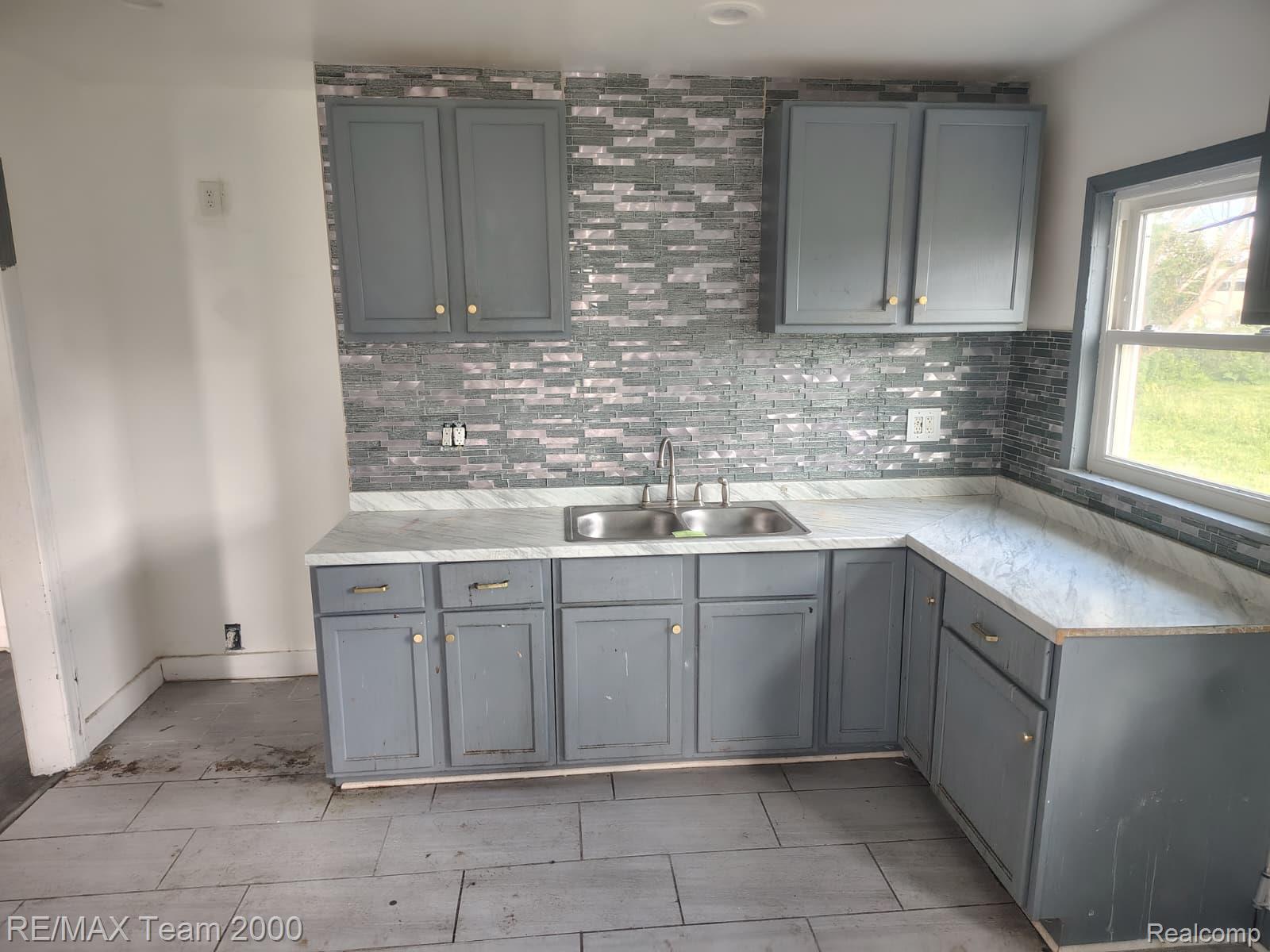8860 Appoline Street Detroit, MI 48228 - Photo 8 of 45 Kitchen featuring L-shaped gray cabinetry with gold-tone hardware, a stainless steel double basin sink, and a multi-tone tile backsplash