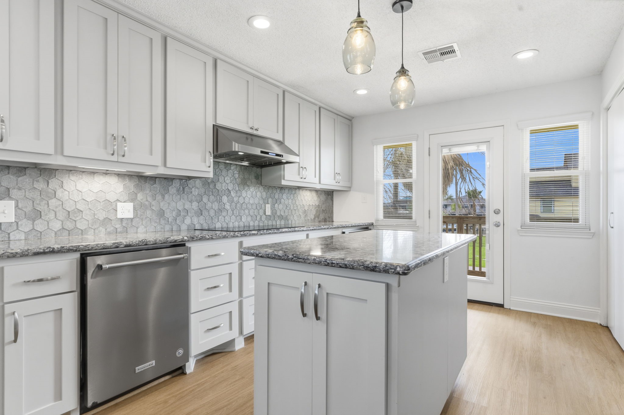 119 Sand Shoals Road Freeport, TX 77541 - Photo 18 of 34 Kitchen toward the back deck — KitchenAid stainless dishwasher, gray shaker cabinetry, glass pendant lighting over the island, and a back door leading to the balcony with cargo lift.