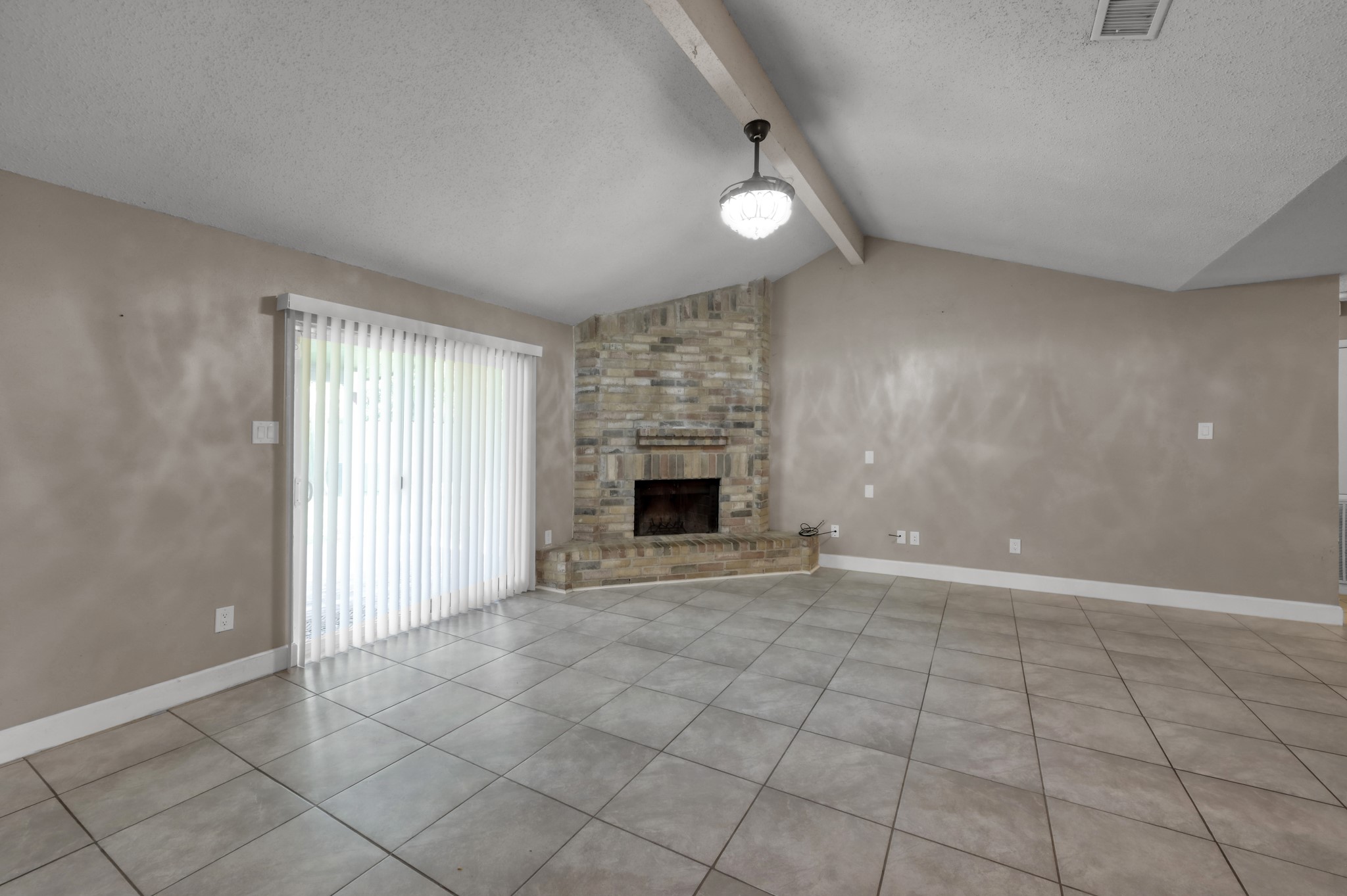 12015 Fallbrook Drive Houston, TX 77065 - Photo 6 of 29 This spacious living room features a high ceiling with an exposed beam, a cozy brick fireplace, large tile flooring, and sliding glass doors with vertical blinds leading to the outside.