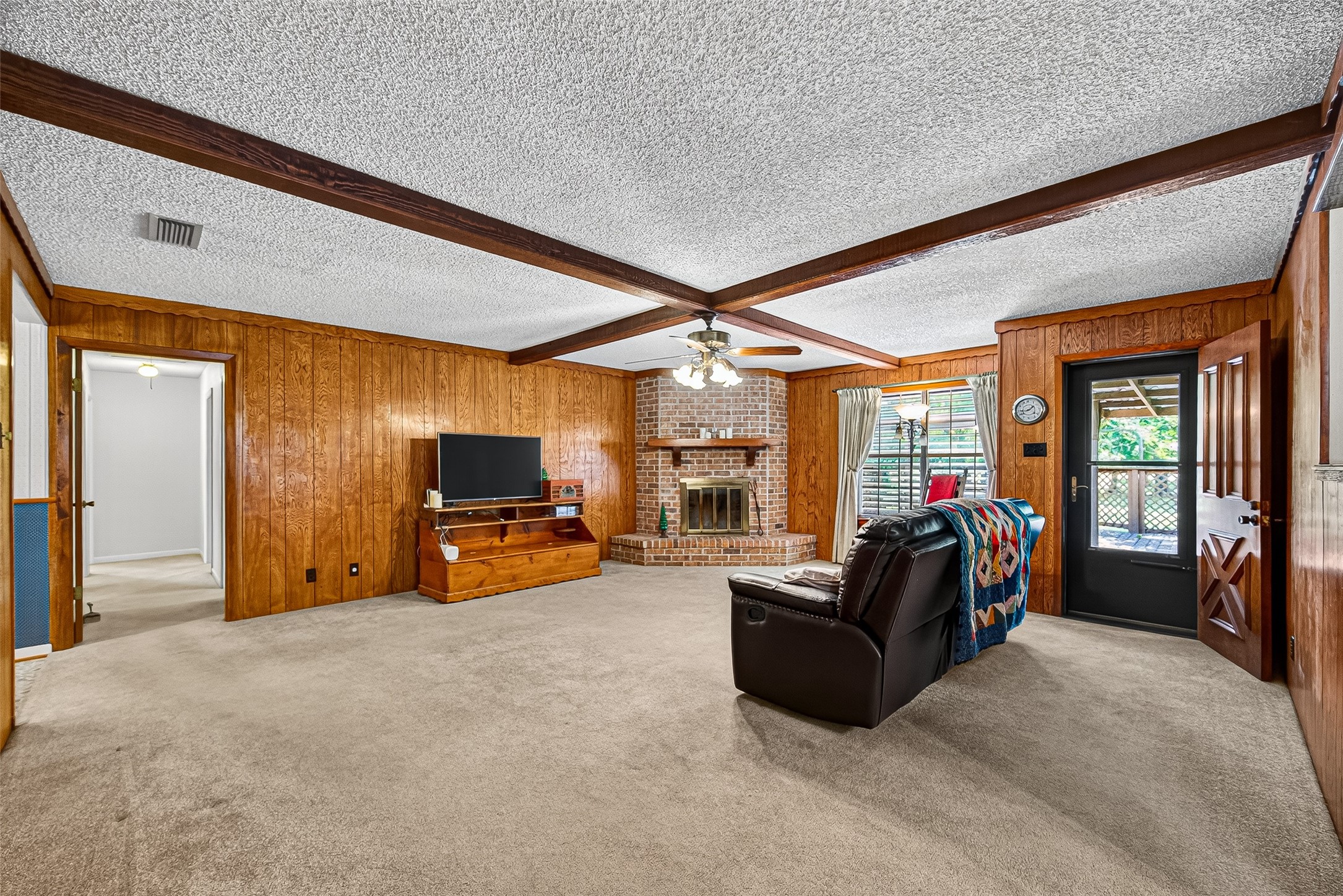 12021 7th 1/2 Street Santa Fe, TX 77510 - Photo 15 of 49 This cozy living room features wood-paneled walls, a brick fireplace, and a ceiling with exposed beams. It has carpeted flooring, a ceiling fan, and large windows allowing natural light. A door leads to an outdoor space.