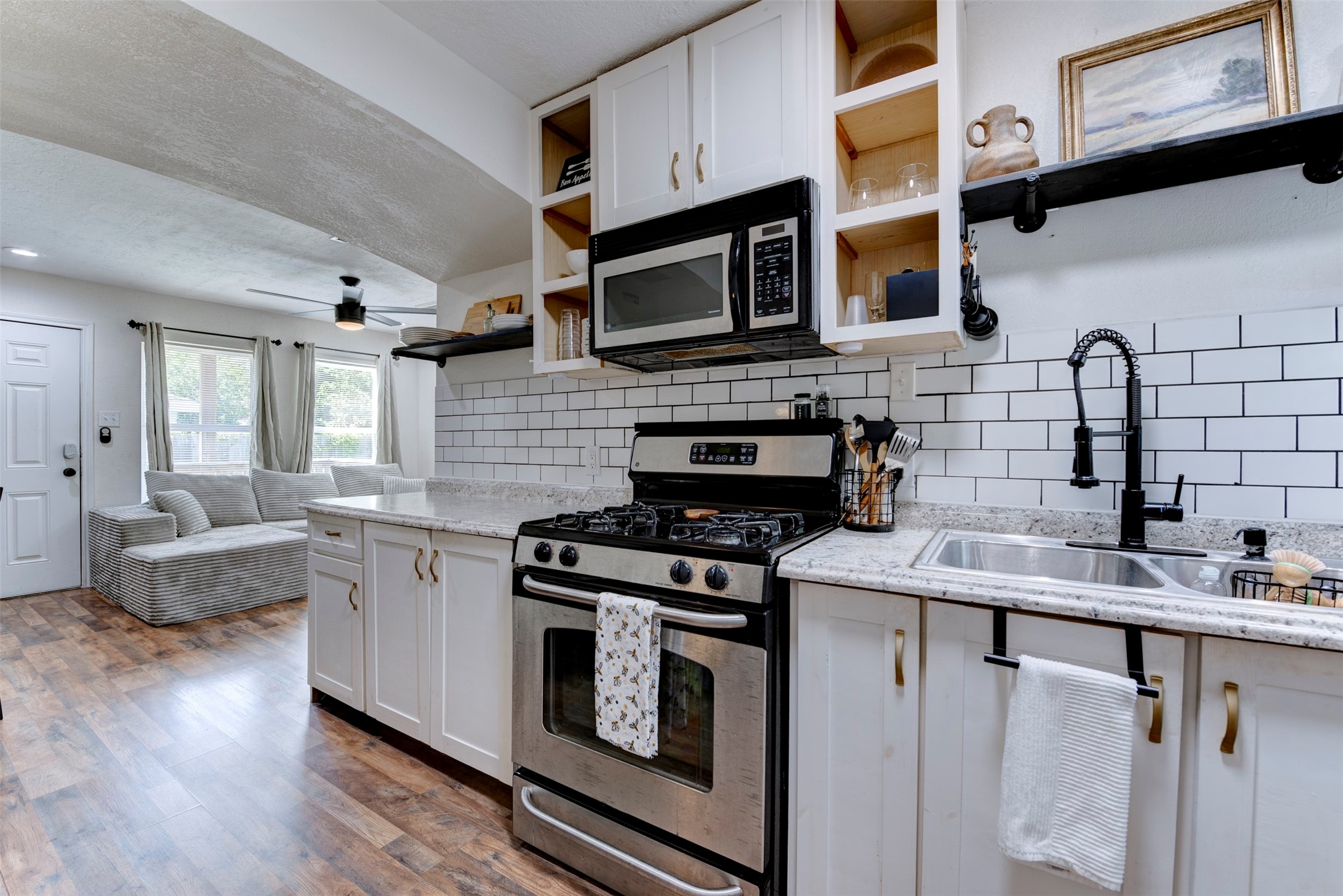 1121 Panama Street Houston, TX 77009 - Photo 10 of 35 Kitchen to living room view showcasing GE gas range, stainless over-the-range microwave, matte black pull-down spring faucet, double-basin stainless sink, granite countertops, white subway tile backsplash with dark grout, white shaker cabinetry with brushed gold hardware, open pine cubbies, black pipe bracket floating shelf, dark wood-look LVP flooring, ceiling fan with light kit, and arched opening to living room.