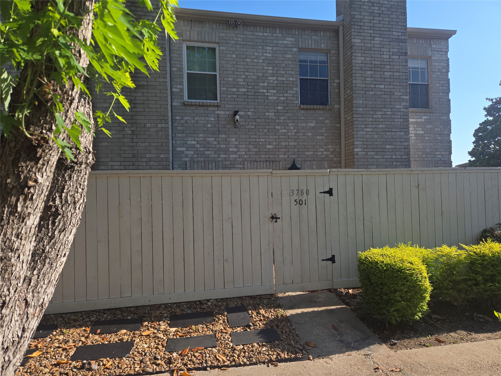 3780 Tanglewilde Street, Unit 501 Houston, TX 77063 - Photo 24 of 26 This photo shows a two-story brick home with a large chimney, fenced yard, and well-maintained shrubs. There's a path leading to a gate, providing privacy and curb appeal.