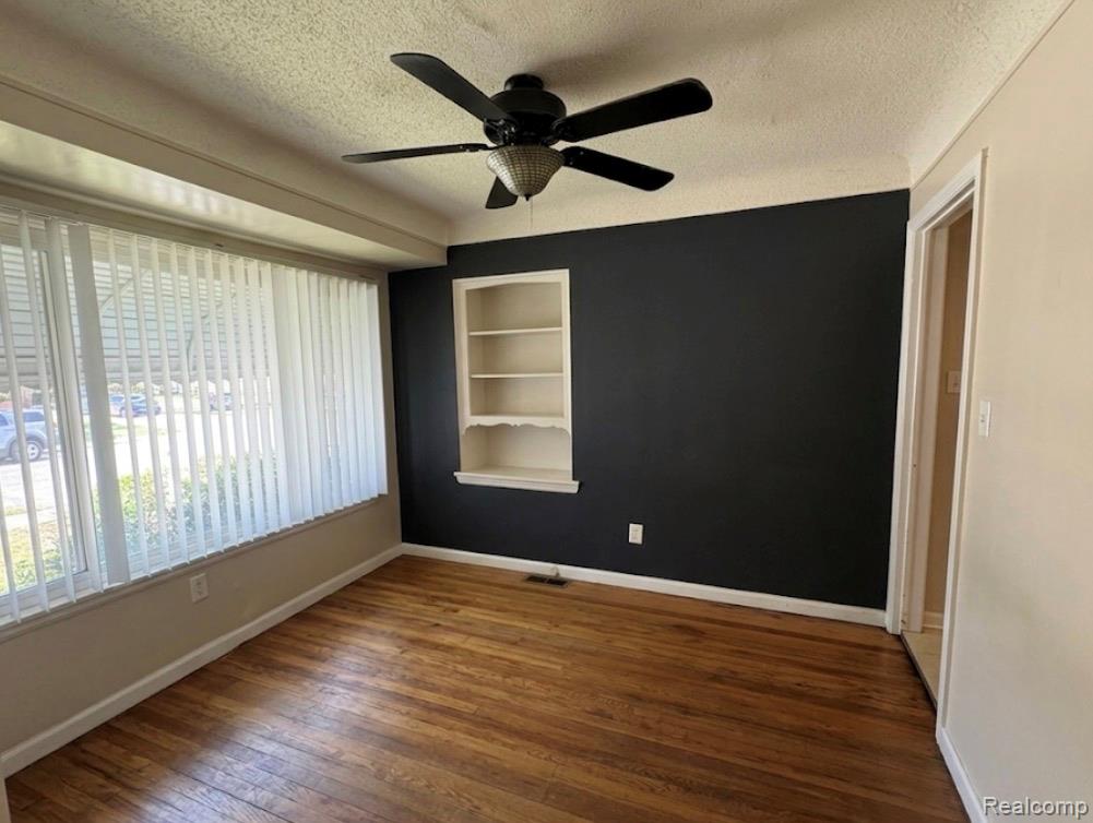 Empty room with a textured ceiling, built in shelves, dark wood-type flooring, and ceiling fan