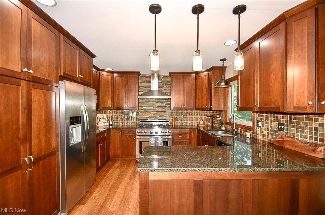 781 Yager Road Clinton, OH 44216 - Photo 19 of 34 Kitchen with decorative light fixtures, backsplash, brown cabinets, wall chimney range hood, light hardwood floors, appliances with stainless steel finishes, and dark stone counters
