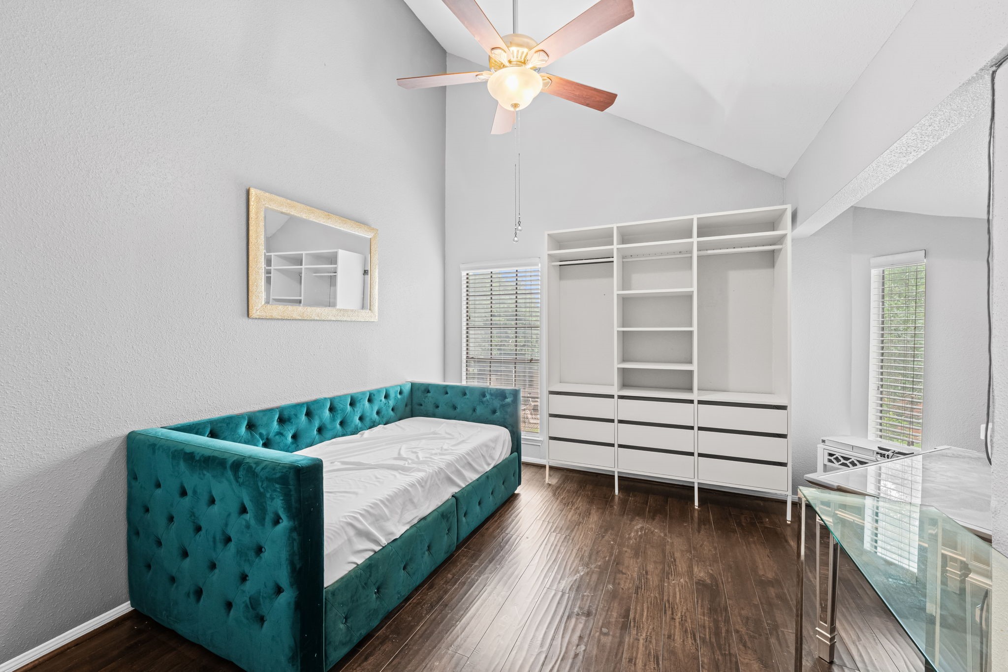 2211 South Kirkwood Road, Unit 1 Houston, TX 77077 - Photo 13 of 16 The secondary bedroom has a cathedral-height vaulted ceiling that rises well above what any standard second-floor room would offer. A ceiling fan sits at the apex, two windows on adjacent walls bring natural light from both directions, and a full-wall built-in shelving and wardrobe system in white provides substantial organized storage without consuming floor space. It's a room that earns its footprint.
