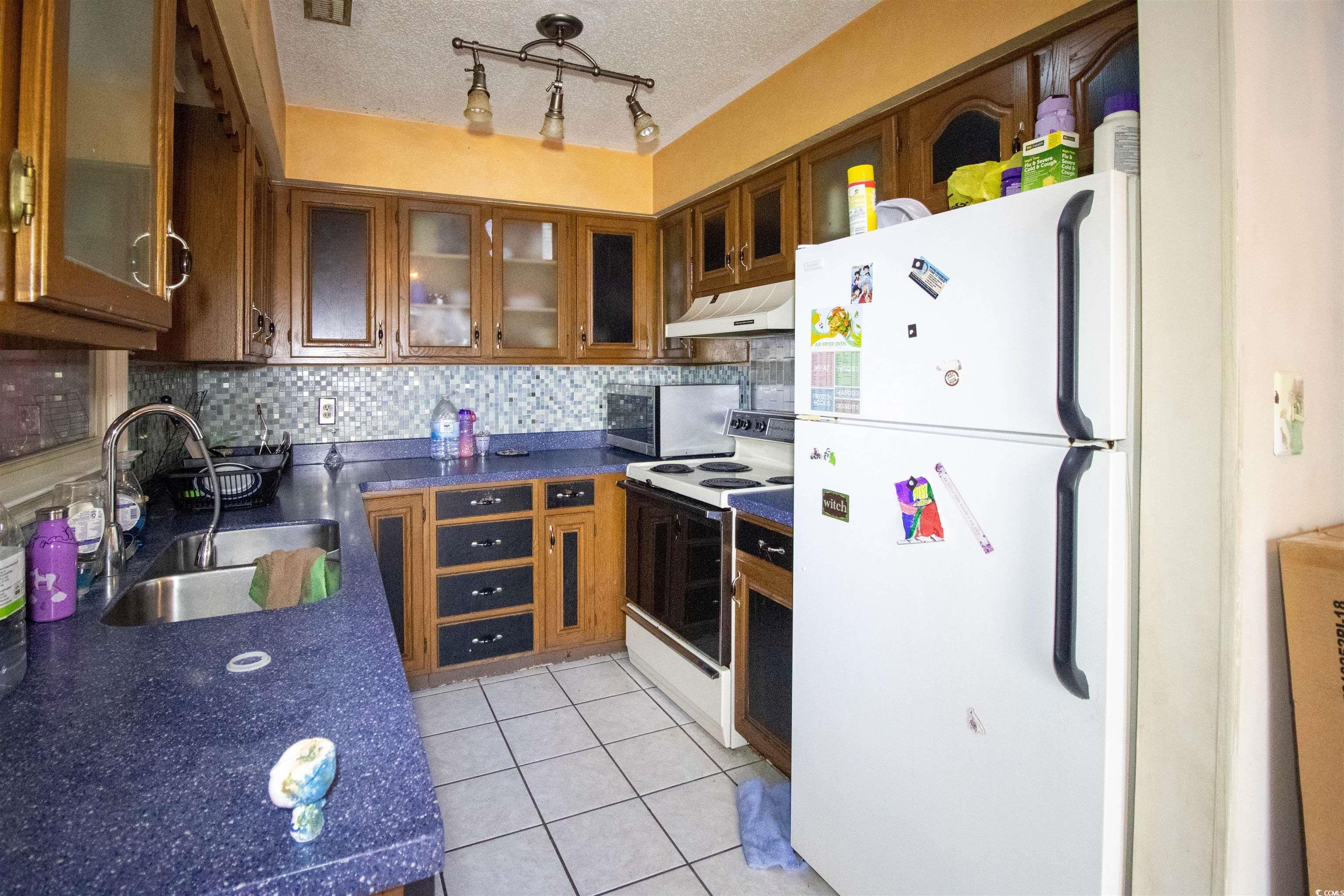 41 Channing Drive Little River, SC 29566 - Photo 5 of 19 Kitchen featuring white appliances, light tile patterned floors, brown cabinetry, under cabinet range hood, and tasteful backsplash