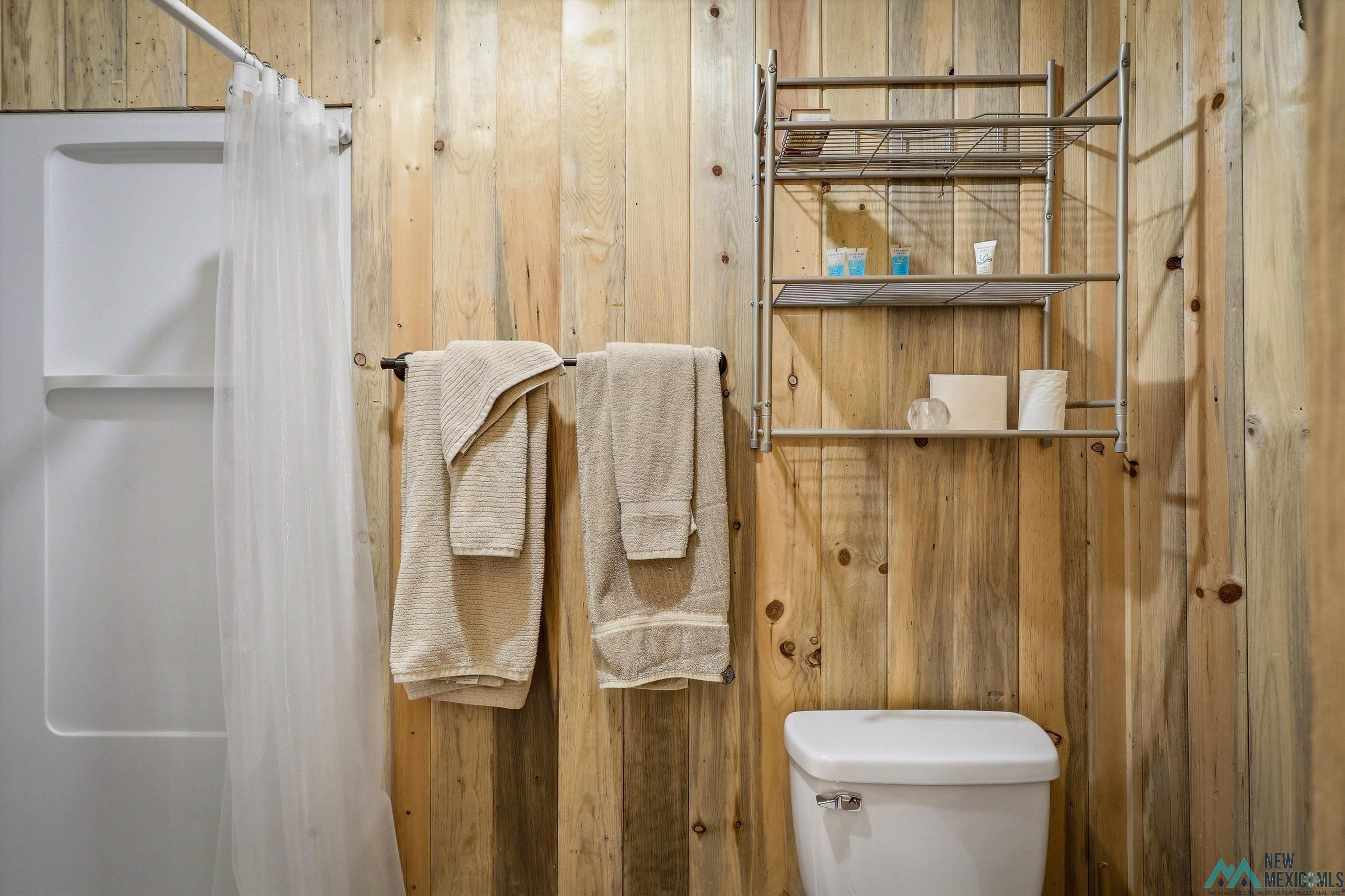 83 Juniper Road Chama, NM 87520 - Photo 38 of 56 Vertical wood-finish wall paneling, white shower surround, white toilet, metal wall-mounted shelving, and dark-finish towel bar