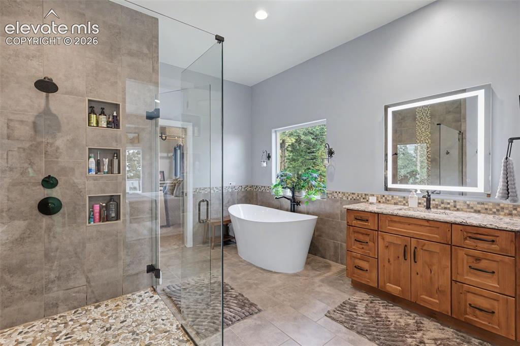21 Leaming Road Colorado Springs, CO 80906 - Photo 29 of 48 Spacious bathroom featuring a freestanding soaking tub, a large walk-in shower with pebble tile flooring and built-in niches, a light wood-finish vanity with a stone countertop, and an illuminated mirror