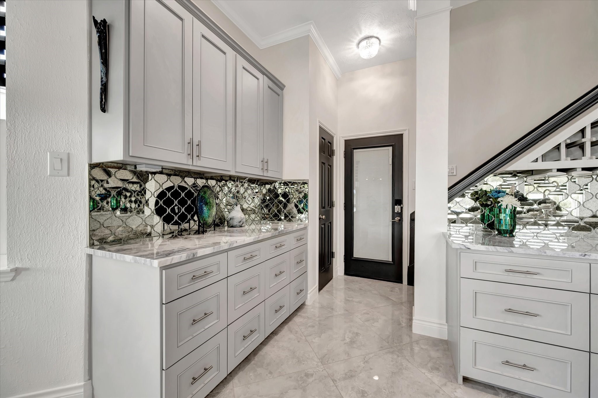 2847 Everett Drive Friendswood, TX 77546 - Photo 24 of 50 This photo showcases a modern hallway with elegant cabinetry and a striking mosaic tile backsplash. The space features marble countertops, light gray cabinets, and glossy tiled flooring. A black-framed door and a staircase with a decorative railing add sophistication.