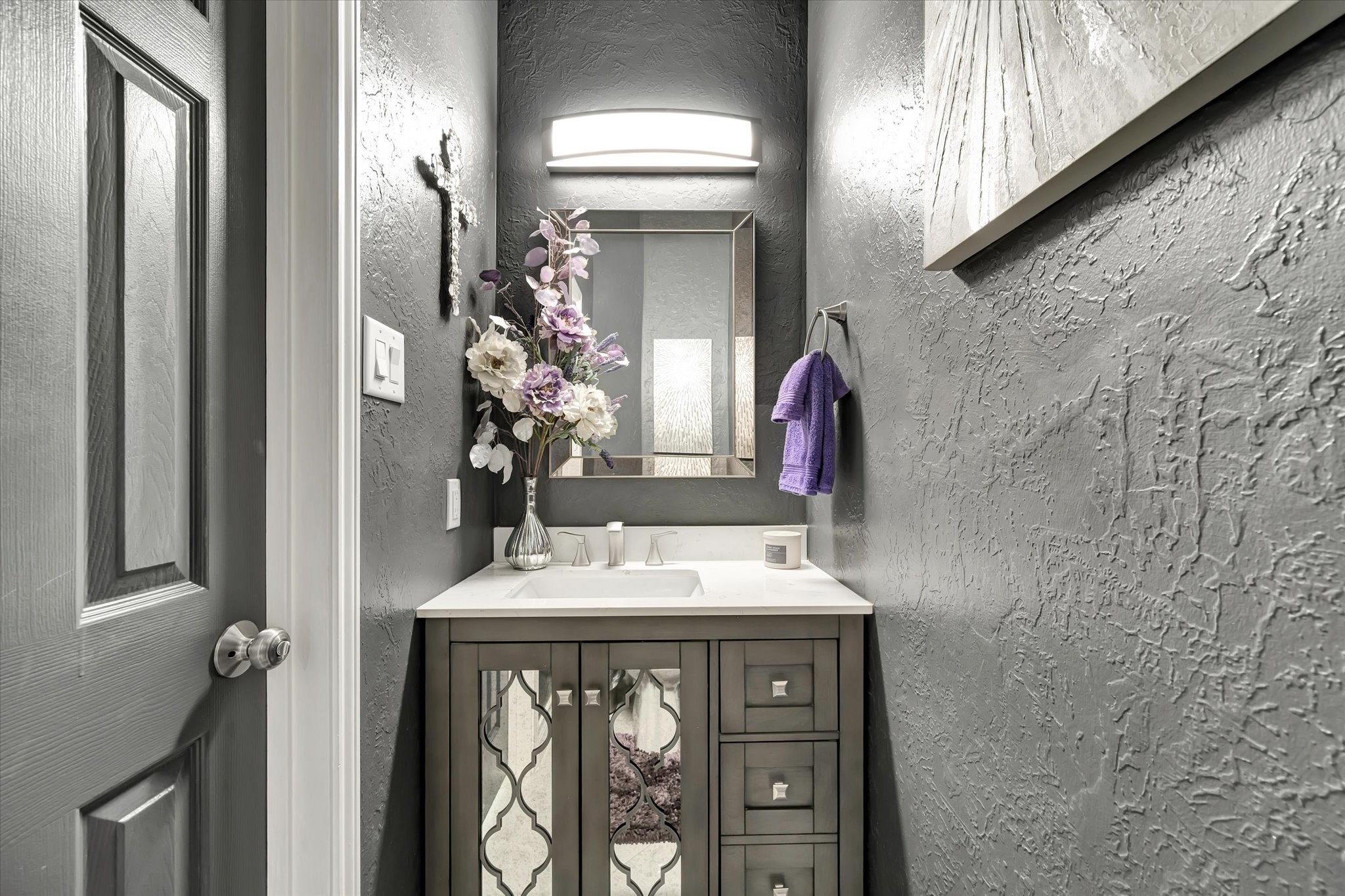 2847 Everett Drive Friendswood, TX 77546 - Photo 28 of 50 This photo shows a stylish powder room with textured gray walls, a modern vanity with decorative cabinet doors, and a well-lit mirror.