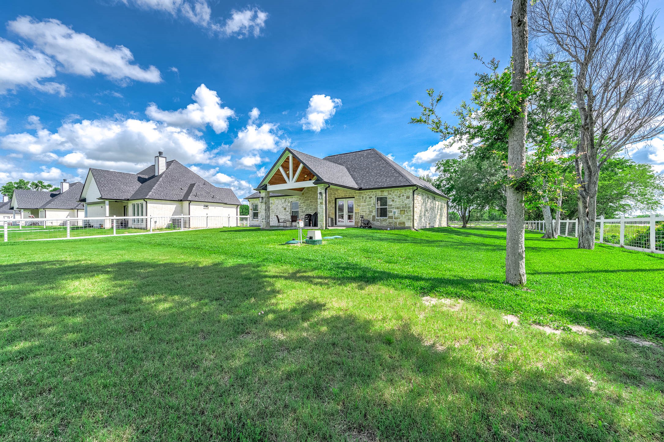 8032 Short Road Needville, TX 77461 - Photo 31 of 50 This view highlights the home’s beautiful stone exterior, spacious yard, and peaceful setting surrounded by open green space and mature trees. The covered patio and fenced backyard add to the property’s charm and outdoor appeal.