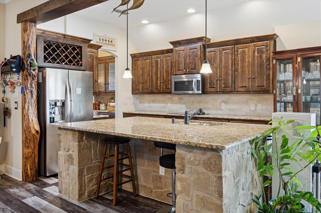 119 George Owens Road Ponder, TX 76259 - Photo 9 of 26 Kitchen featuring an expansive stone-clad island with a sink, pendant lighting, wood-finish cabinetry, a tile backsplash, and stainless steel appliances