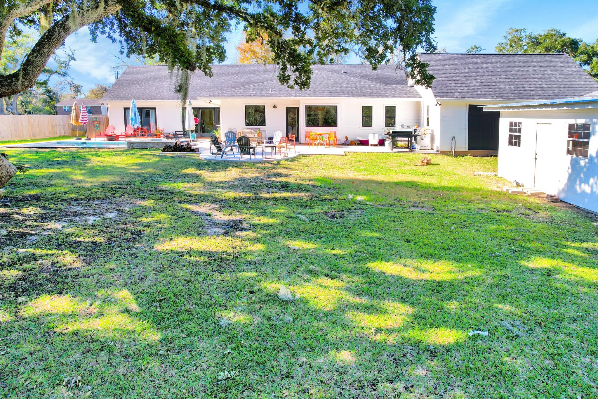 295 County Road 687 Angleton, TX 77515 - Photo 11 of 38 This photo showcases a spacious backyard featuring a well-maintained lawn, a patio area with outdoor seating, and shaded trees. The house has a modern exterior with large windows, and there's an additional structure on the side, possibly a shed or garage. Ideal for outdoor entertaining and enjoying open space.
