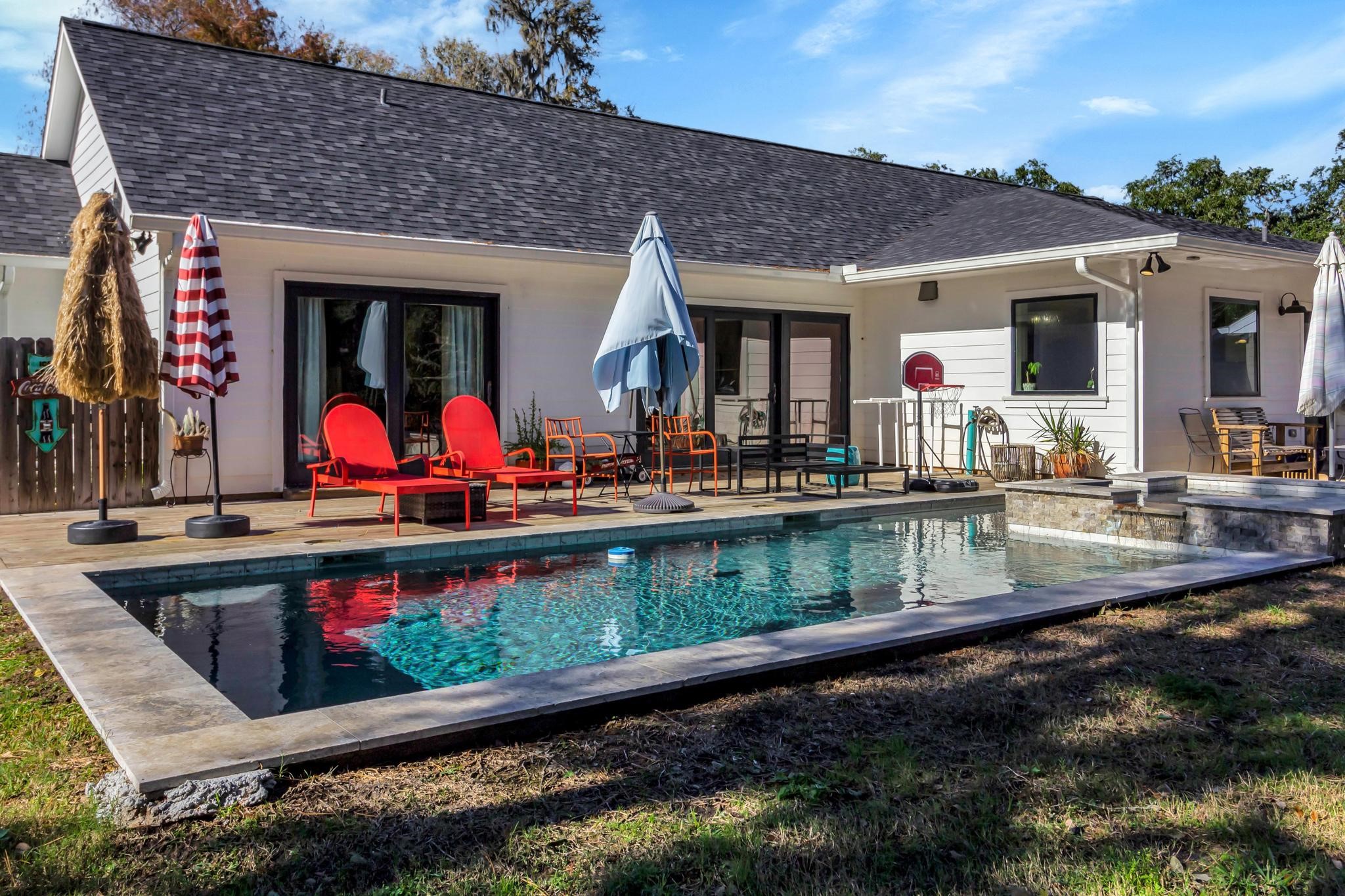 295 County Road 687 Angleton, TX 77515 - Photo 9 of 38 This backyard features a modern patio with red lounge chairs, umbrellas, and a sleek rectangular pool. The house showcases white siding and large sliding doors, creating an inviting outdoor living space.