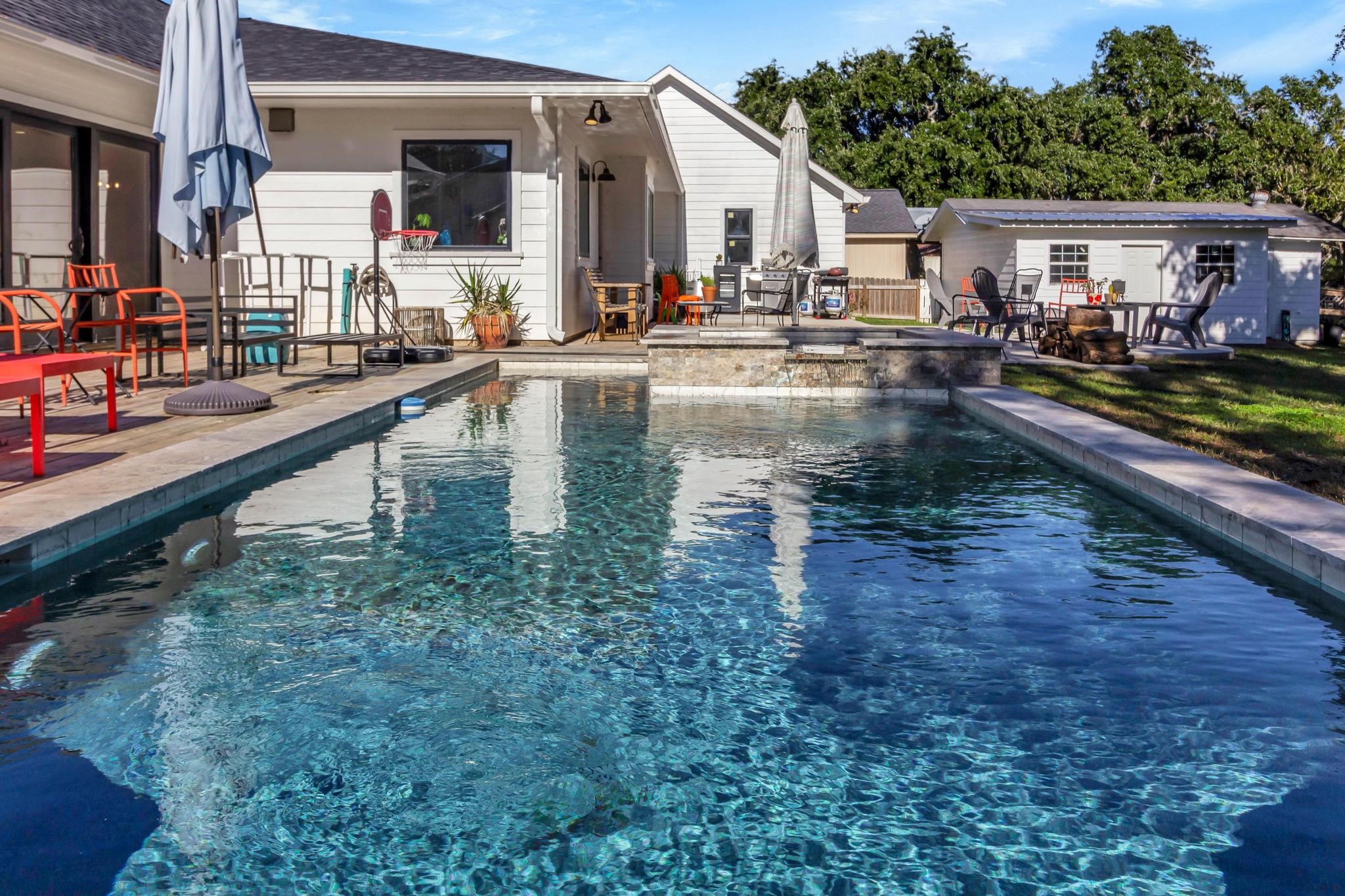 295 County Road 687 Angleton, TX 77515 - Photo 10 of 38 This photo showcases a charming backyard with a sparkling pool, surrounded by a spacious patio area perfect for relaxation and entertaining. The house features a modern exterior with large windows, and there's additional seating and a finished and air conditioned outbuilding in the background, all set against a backdrop of lush greenery.