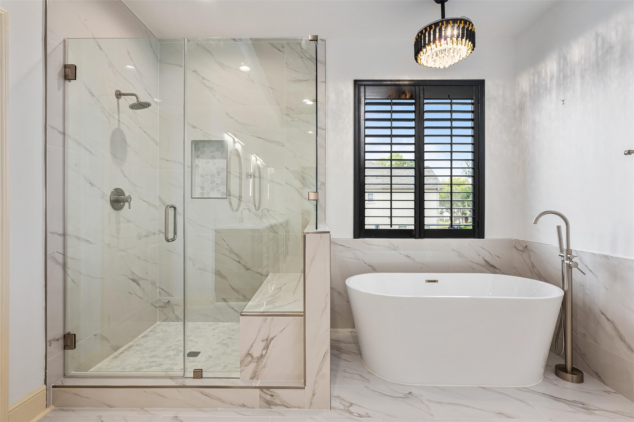 4523 Maple Street Bellaire, TX 77401 - Photo 28 of 50 The walk-in shower features a frameless glass enclosure and a marbled tile surround that extends to the ceiling, complete with a bench seat and accent tiles along the floor and wall niche. Adjacent to the shower, a freestanding soaking tub with an upgraded faucet and chandelier offers a spa-like retreat, while a window with plantation shutters provides natural light and privacy.