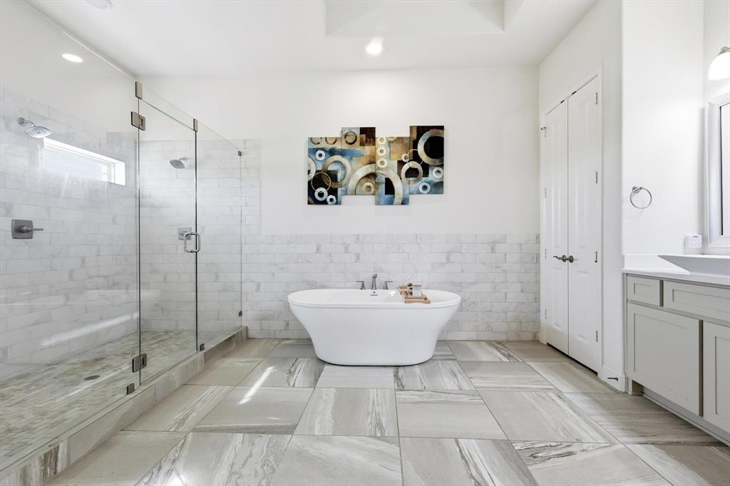 7751 Faught Road Northlake, TX 76226 - Photo 21 of 39 Modern bathroom featuring a freestanding soaking tub, a frameless glass shower enclosure with dual showerheads, light gray tile flooring, a single vanity with an undermount sink, and white shaker-style cabinetry