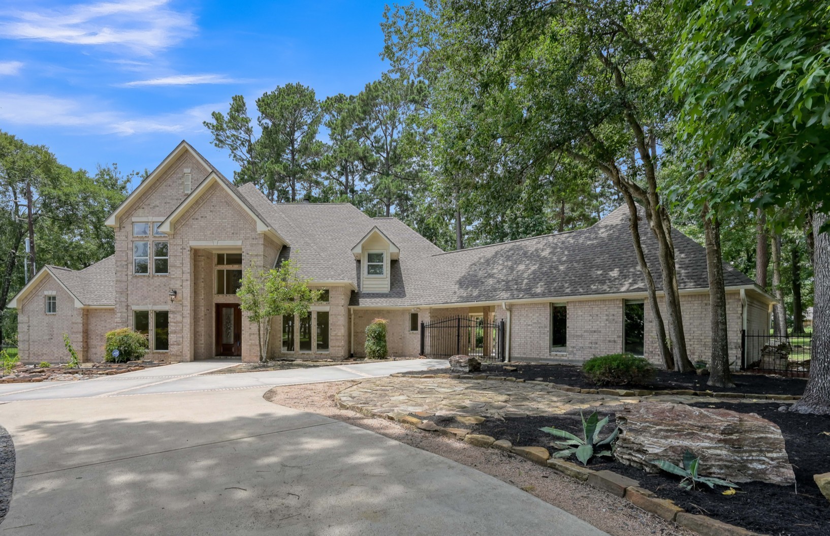 This photo showcases a spacious, elegant home with a light brick exterior and a gabled roof. The house is set amidst a lush, wooded landscape, featuring a wide driveway and well-maintained landscaping, creating a welcoming and serene atmosphere.
