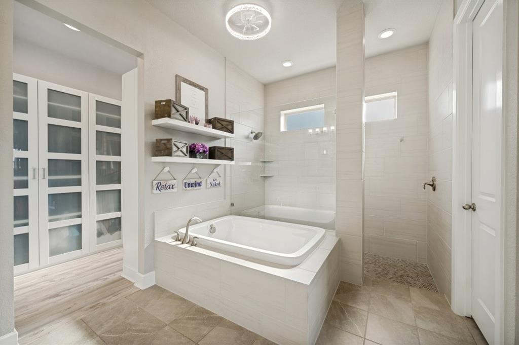 2721 Old Shire Path Road Prosper, TX 75078 - Photo 11 of 39 Spacious bathroom featuring a built-in soaking tub with brushed nickel fixtures, a walk-in shower with pebble flooring and tiled walls, a frosted glass privacy window, and a separate room with built-in cabinetry and glass-front doors